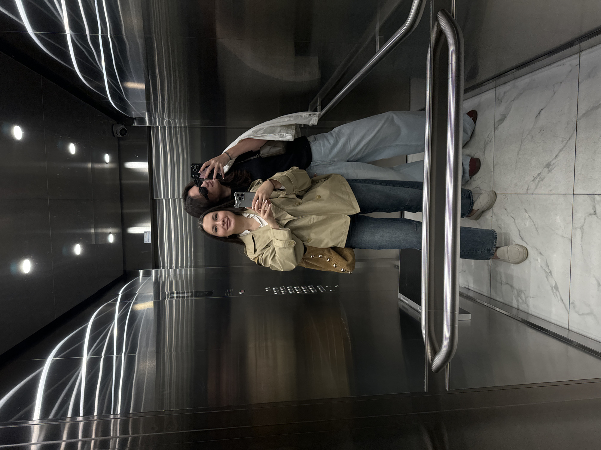 Two women are posing in an elevator. They are standing next to each other, and each is holding a smartphone to take a picture of themselves in the mirror. The woman in the front is wearing a tan trench coat, jeans, and off-white sneakers, and the other is wearing a black top, a white jacket, jeans, and red sneakers. The elevator has a shiny metal interior with illuminated lines on the wall, and a panel of buttons. The flooring is made of white marble tiles. There is a handrail on the side. The scene is likely in Belgrade, Serbia.