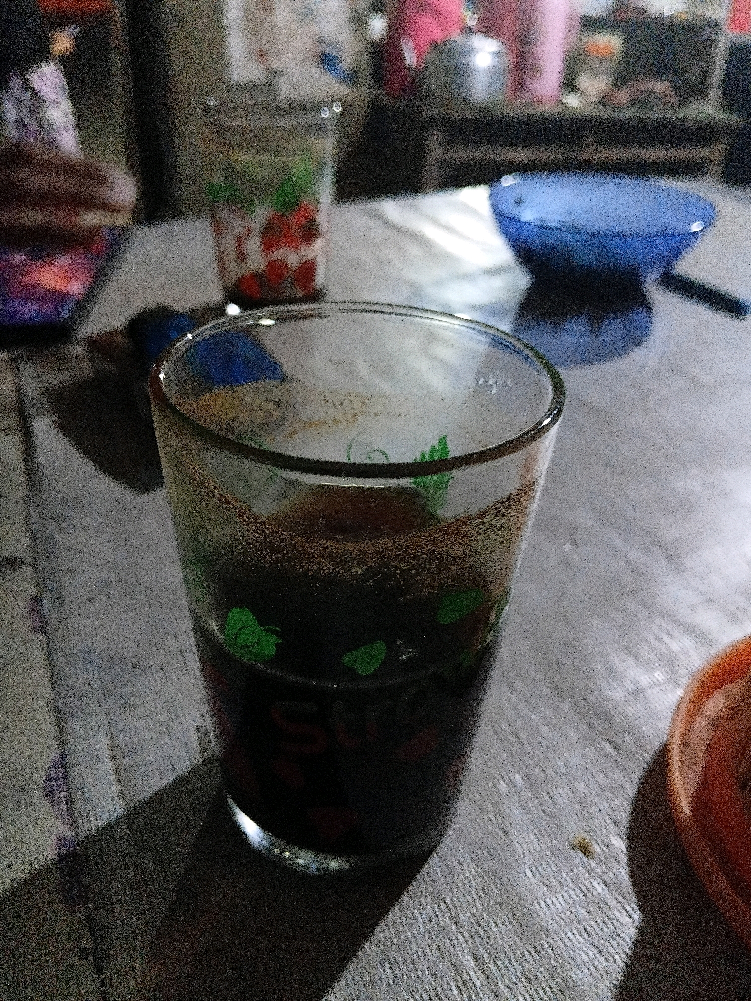 The image captures a close-up view of a glass of dark liquid, likely coffee or tea, with some sediment at the bottom. The glass has a decorative pattern of green leaves and red strawberries. The glass is placed on a textured surface, possibly a wooden table, which is lit by a dim, ambient light source, creating shadows. In the background, other objects are visible but out of focus, including another glass with a similar pattern, a blue bowl, and what appears to be kitchenware. The overall setting suggests a casual indoor environment, potentially a home or a small eatery in Bantarbarang, Indonesia. There are no discernible people or specific actions in the foreground of the image.