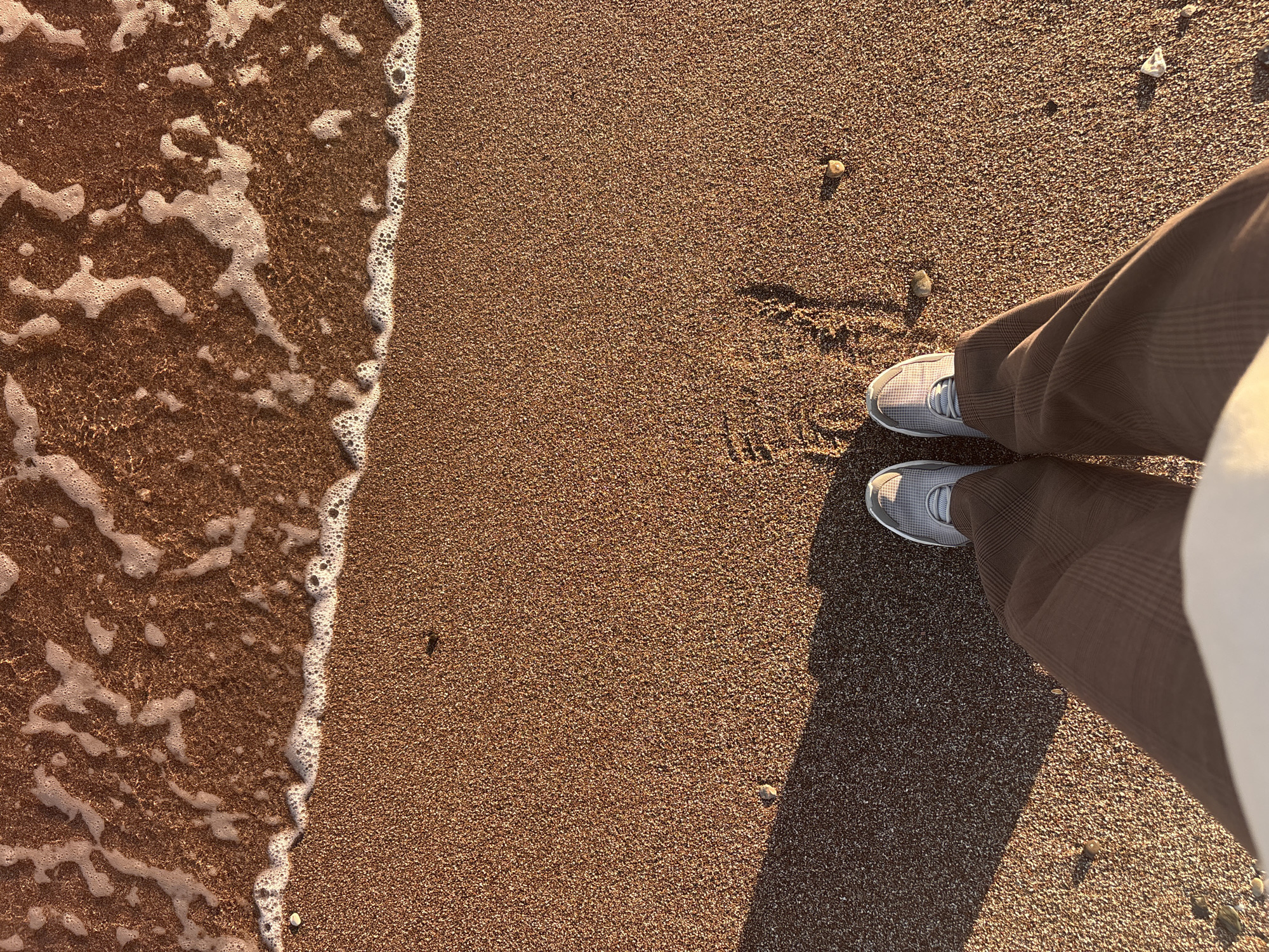 An overhead photograph captures a person's legs and feet standing on a reddish-brown sandy beach. The individual wears brown trousers with a subtle plaid pattern and light grey sneakers with white laces. Their body casts a long, distinct shadow across the sand towards the lower right, indicating a low sun angle, likely early morning or late afternoon.
To the left, foamy white waves gently wash ashore, creating an organic boundary with the dry sand. The sand is densely packed with fine granules and scattered small pebbles or rocks. Faint marks or shallow impressions are visible in the sand near the feet, suggesting recent movement or standing.
The scene depicts a peaceful beach environment under clear, sunny weather, consistent with a coastal location such as Boreti, Montenegro. There is no visible text in the image.