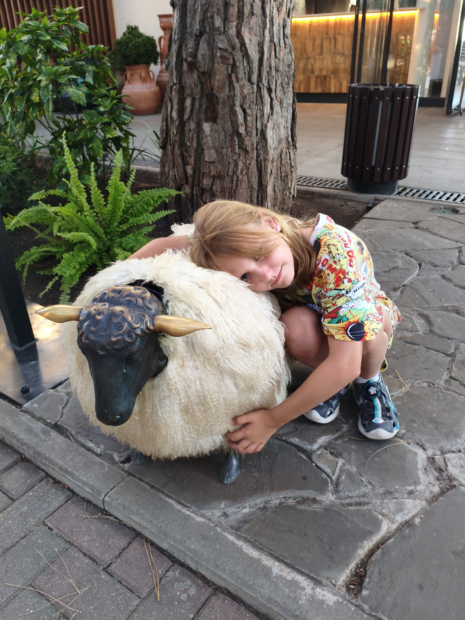 A young girl with blonde hair is crouching next to a sculpture of a sheep. The sheep sculpture is white and fluffy with a dark head and golden horns. The girl is leaning her head against the sheep and smiling at the camera. She is wearing a colorful t-shirt with a comic book-style print and dark sneakers with blue accents.
The scene appears to be outdoors, with a paved walkway made of large, irregular stones. To the left of the girl, there is a patch of green foliage and a large tree trunk. In the background, a modern building with warm lighting is visible, along with a dark wooden trash can. The lighting suggests it is daytime, possibly late afternoon or early evening, given the warm glow from the building. The overall impression is a relaxed and playful moment captured in an outdoor public space. The location context is Gelendzhik, Russia.