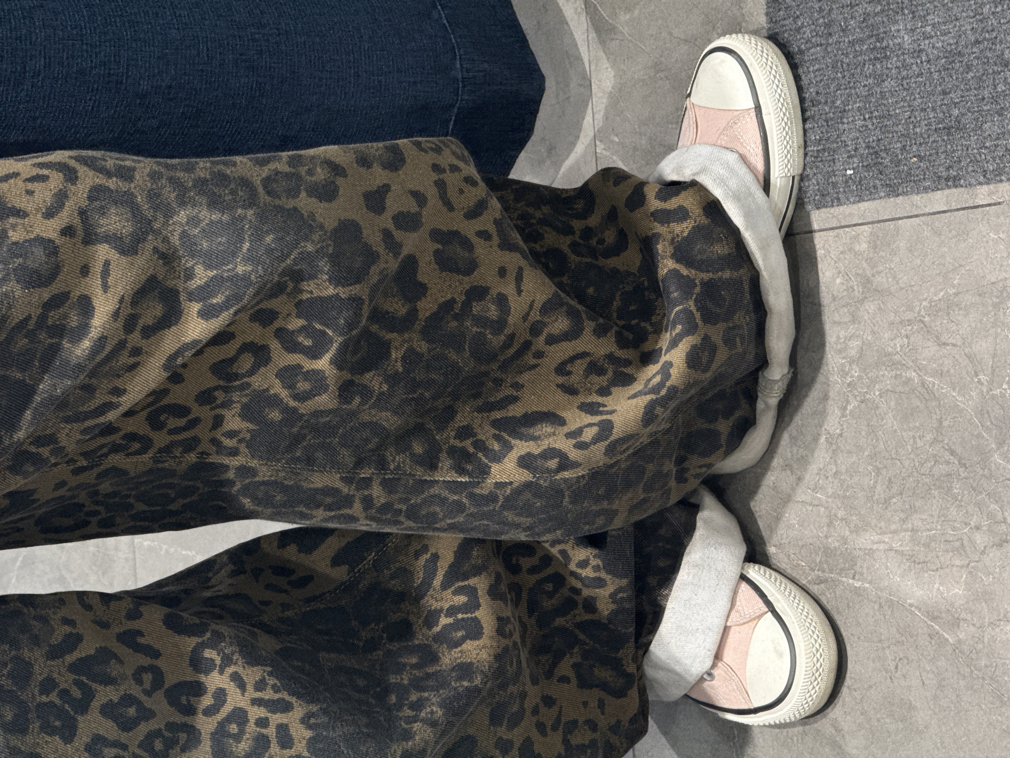 The image shows a person's legs and feet standing on a grey, marble-like tiled surface. The person is wearing wide-legged, dark brown/olive green pants with a dark animal print. The pants are cuffed at the ankles, revealing white socks and pink and white sneakers. A section of dark blue fabric is visible above the pants, suggesting another piece of clothing. No specific details indicate the location is Yerevan, Armenia.