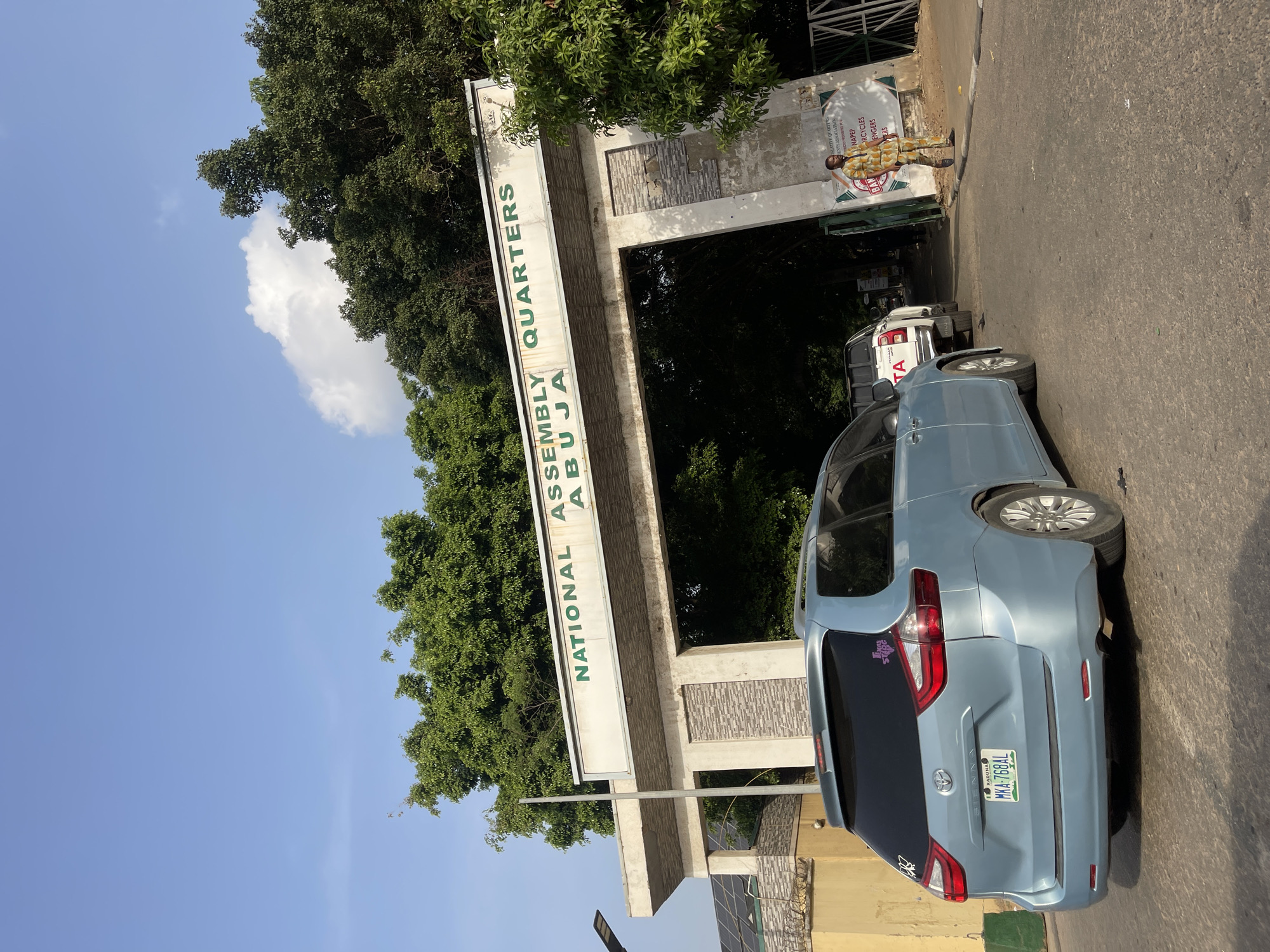 The image captures an outdoor scene at the entrance of a residential area under a bright, sunny sky with scattered clouds. The primary subject is a large, light-colored archway serving as a gate, bearing the inscription "NATIONAL ASSEMBLY QUARTERS ABUJA" in green lettering. Lush green trees are visible behind and around the gate, suggesting a verdant environment.
Several vehicles are present on the paved road in front of the gate. A light blue Toyota Sienna minivan is prominently parked in the foreground, facing away from the viewer, with its license plate "MKA-768AL" from Kaduna, Nigeria, clearly visible. Another white vehicle, possibly a Toyota pickup truck, is parked further down the road behind the minivan.
To the right of the archway, partially obscured, is a banner featuring a standee of a person in uniform. Text on this banner, though partly cut off, includes phrases like "LEGISLATIVE QUARTERS", "ASSOCIATION", "CYCLES", "ENGERS", and "BAN". The overall impression is of a controlled entry point to a significant residential complex in Abuja, Nigeria, during daylight hours. The road appears to be made of asphalt.