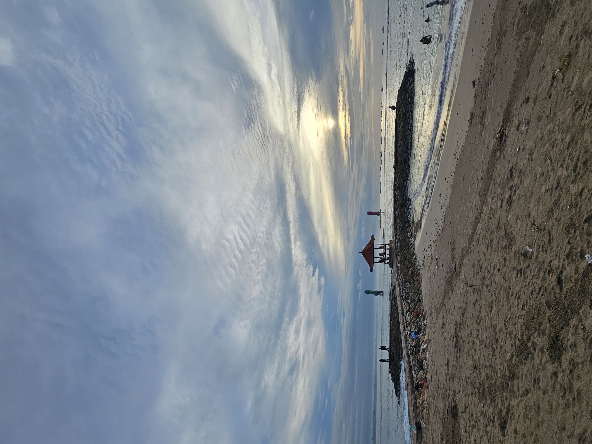 The image captures a coastal scene at Denpasar, Indonesia. The sky, occupying a significant portion of the frame, is filled with streaky clouds partially obscuring the sun. The shoreline is lined with rocks, and gentle waves are visible. A red pavilion stands on the beach. A green marker is nearby. Figures of people are distributed along the shoreline. The sand, coarse and uneven, covers the lower right corner of the image, with some debris scattered on it.