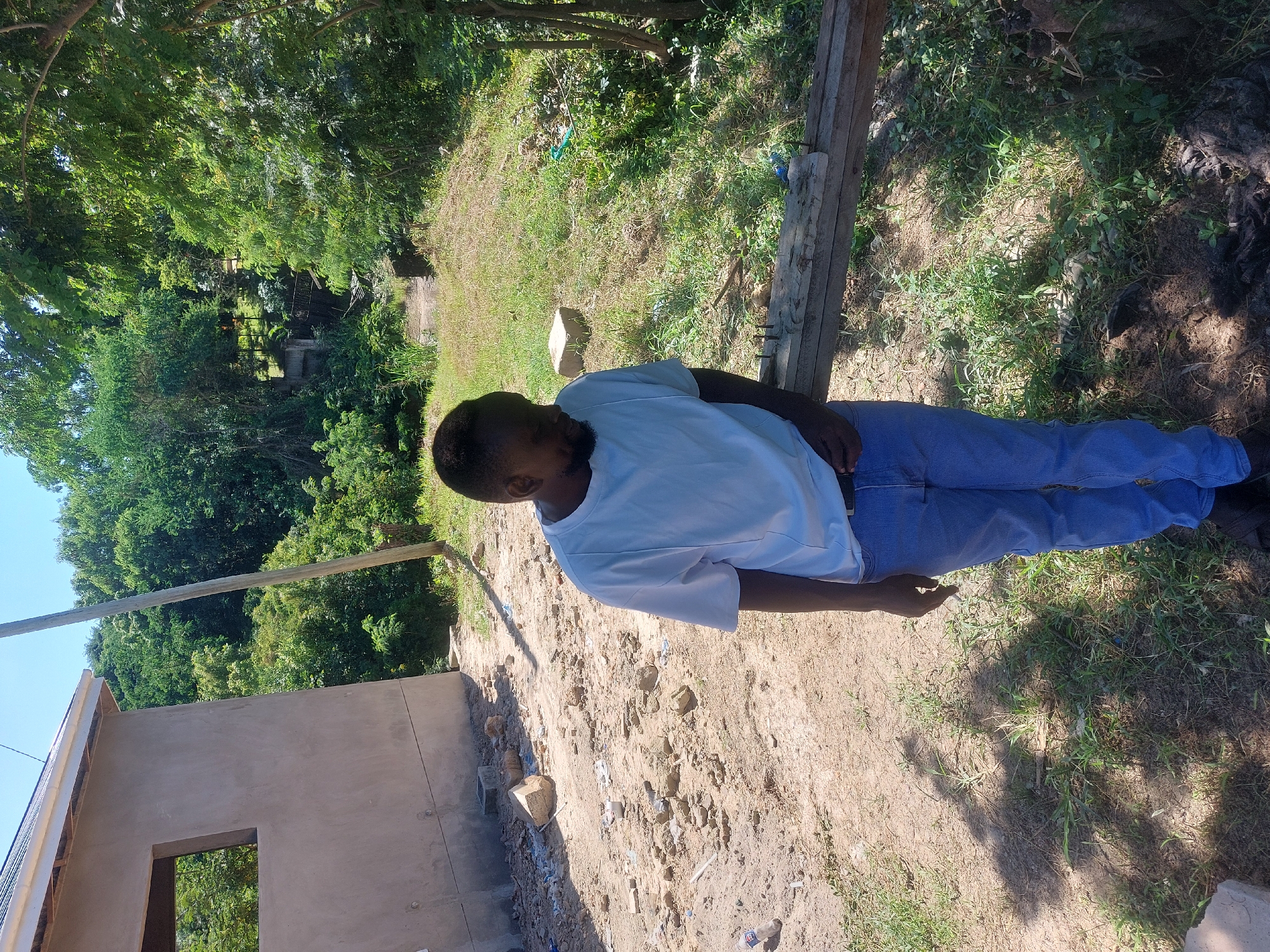 The image shows a man in Dar es-Salaam, Tanzania. He is wearing a white t-shirt and blue jeans, standing on a dirt ground. To his left is a partially constructed building with a window opening. The scene is outdoors, with trees and vegetation in the background, suggesting a peri-urban environment. There are wooden beams and other construction materials nearby. The lighting indicates a sunny day. The area appears to be a construction site or a property under development.