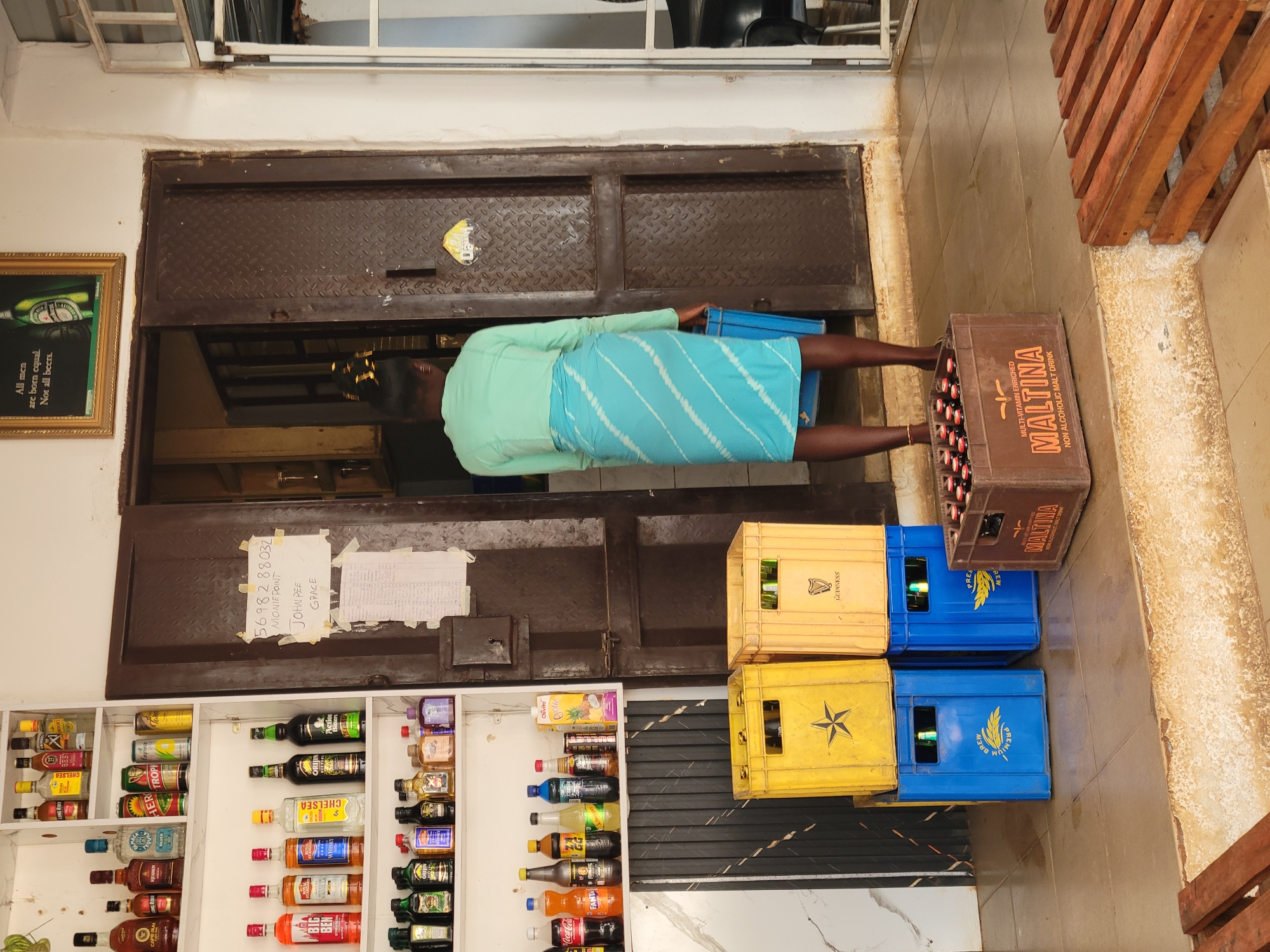 The image presents an indoor scene, likely a small provision shop or bar, with a person actively engaged in handling merchandise. A person, viewed from behind, wears a light blue long-sleeved top and a striped blue and white skirt or wrapper. They stand in front of a dark, diamond-patterned metal door, holding a blue crate.
To the person's right, a stack of beverage crates includes a brown crate labeled "MALTINA MULTI-VITAMIN ENRICHED NON ALCOHOLIC MALT DRINK" filled with bottles. Below this are blue crates marked "PREMIUM BREW" and yellow crates with "GUINNESS" branding, also containing bottles.
On the left side of the frame, a white shelving unit is filled with a variety of bottled beverages. These include numerous alcoholic spirits such as "CHELSEA", "LONG RIDE", "HERO TROPHY", "BIG BEN", "ORIJIN", and "Action Bitters", alongside non-alcoholic options like "Coca-Cola", "FANTA", "Chivita", and "Qyotic". Many of the spirit bottles are small, miniature sizes.
Above the shelves, taped to the metal door, are two pieces of paper. One clearly displays text including a phone number "5698288032" and names like "MONIEPOINT JOHAN PEE GRACE". To the far left, a framed picture on the wall features a Heineken bottle and the slogan: "All men are born equal. Not all beers." The floor appears tiled, and a set of wooden slats or pallets are visible on the right.
The overall scene suggests a routine activity of stocking or organizing drinks within a commercial establishment in Barakin Akawo, Nigeria. The lighting indicates an indoor setting, and no specific time of day or weather conditions are discernible.