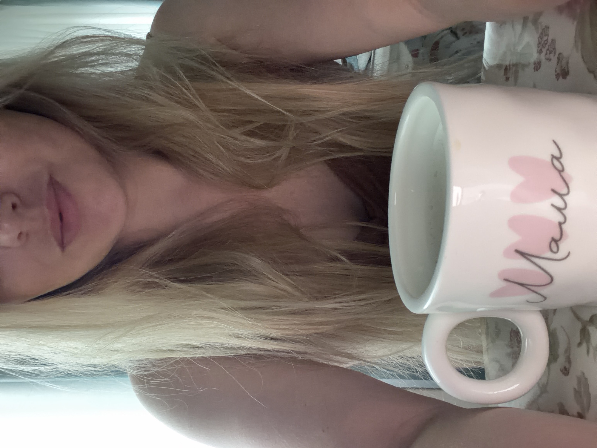 A woman with long, blonde hair is holding a white mug with pink hearts and black cursive writing. The mug appears to be filled with a light-colored liquid. The background is indistinct but includes floral patterns. The location is not discernible from the image.