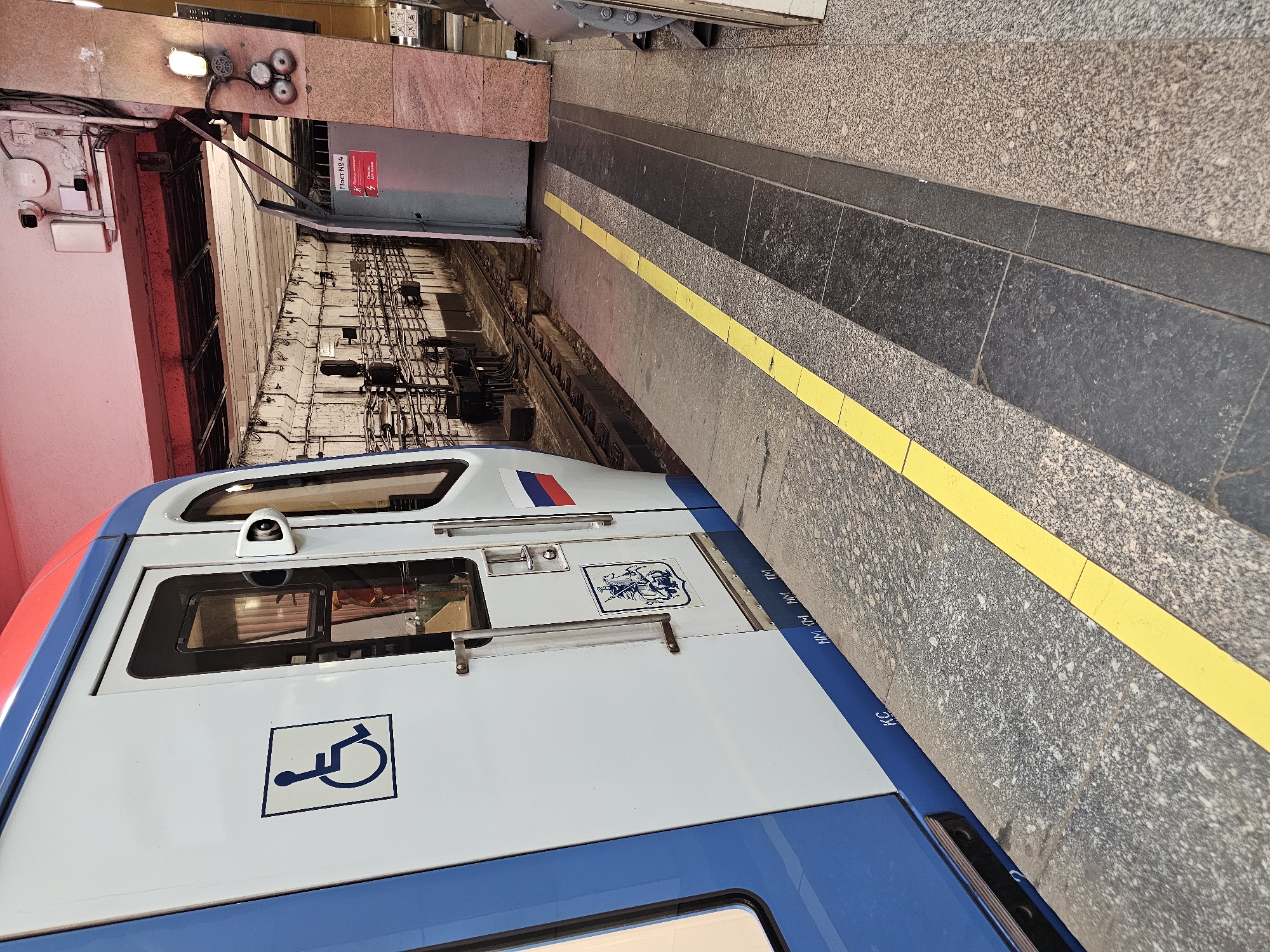 The image shows a subway platform with a train alongside. The train is white with blue and red accents. A blue wheelchair accessibility symbol is visible on the side of the train. The platform is made of granite tiles, with a yellow safety line along the edge. In the background, there are tracks, electrical equipment, and a closed door with Russian text and a caution sign. The scene appears to be indoors, likely in a subway station.