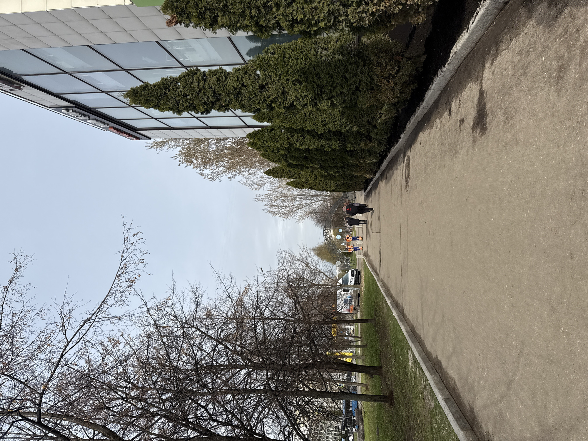 The image shows a view along a sidewalk in Moscow, Russia. A modern building with glass windows and coniferous shrubs borders one side of the sidewalk. On the other side are bare-branched trees and a grassy area. In the distance, several pedestrians are walking on the sidewalk, and orange-vested workers are visible. Cars are parked along the street beyond the trees. The sky is overcast.