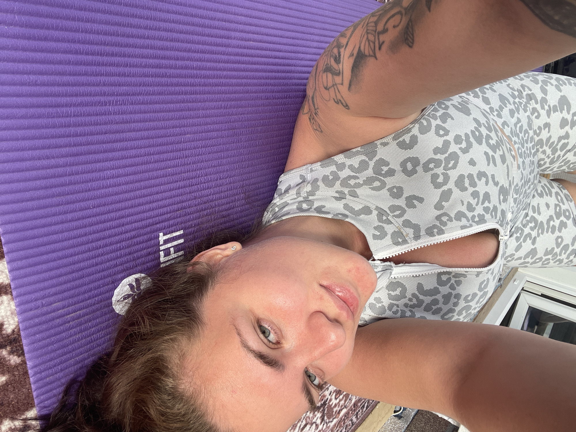 A woman is lying on a purple yoga mat. She is wearing a gray and white patterned athletic outfit. She has a tattoo on her left upper arm. The yoga mat has a white logo on it. There is a window with blinds in the background. The image appears to be a selfie.