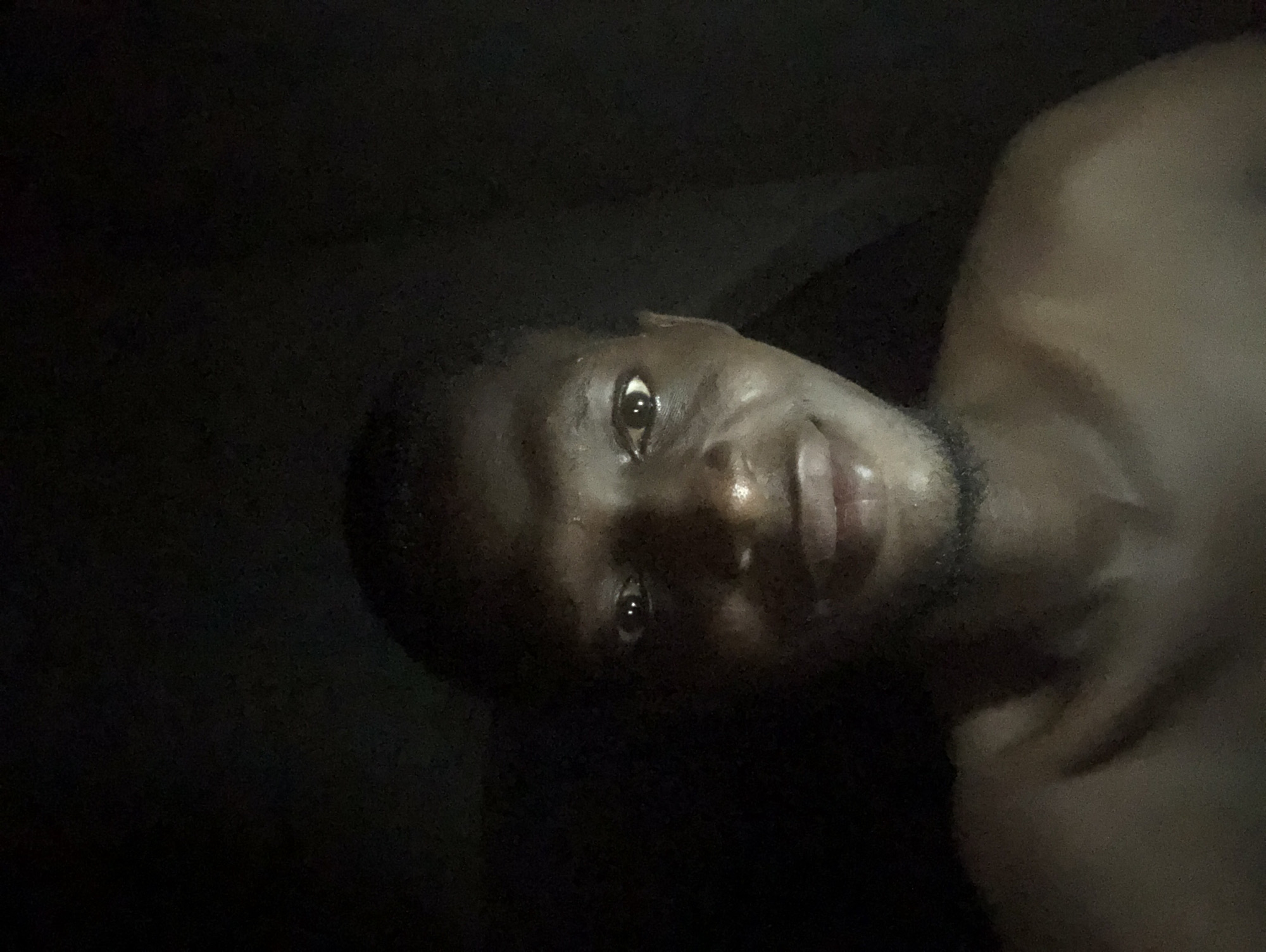 This is a dimly lit, close-up portrait of an individual, likely a young adult male, with dark skin. The image is rotated, presenting the subject's head and upper torso turned almost 90 degrees counter-clockwise from an upright orientation.
The person's face is partially illuminated by a strong, directional light source, creating dramatic highlights and deep shadows. One eye, wide open and reflecting the light intensely, is prominently featured, along with part of the nose, lips, and chin. A faint smile or smirk is visible. Subtle stubble can be seen on the chin and jawline. The other side of the face is largely obscured by darkness, with only a very faint outline of the second eye discernible in the deep shadow.
The individual appears to be shirtless, with bare skin visible on the shoulder and upper chest, which also reflects some of the light and appears somewhat shiny, possibly from sweat or oil. The background is uniformly dark and featureless, offering no specific location cues. The overall mood is intimate and captured in extremely low-light conditions. No visible text is present in the image.
