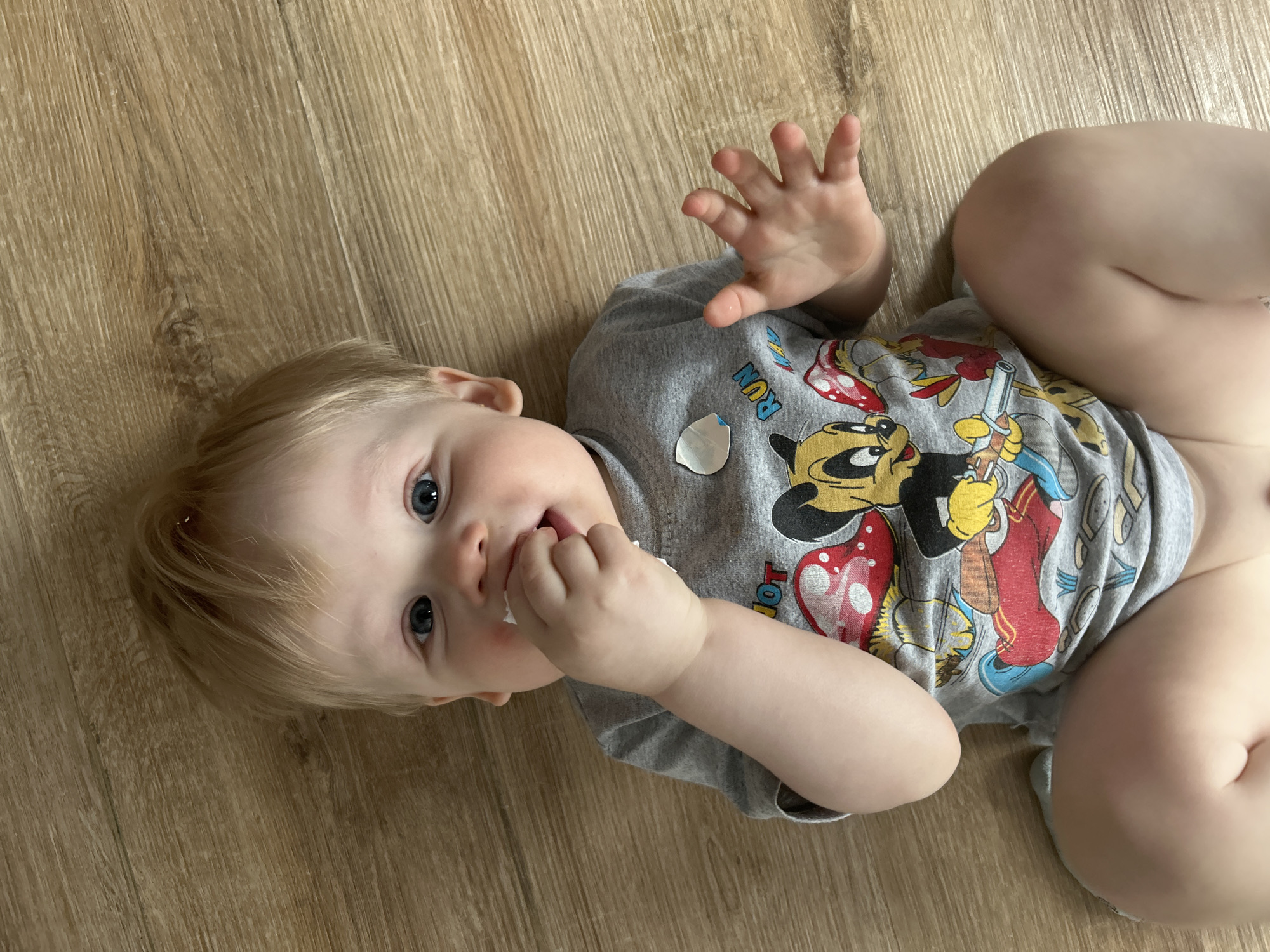 A light-skinned baby with blond hair and blue eyes lies on their back on a light brown wooden floor. The baby is wearing a grey t-shirt featuring colorful illustrations of Disney characters, including Mickey Mouse, Donald Duck, Goofy, and Pluto. Partial text "RUN" and "TOY" are visible on the shirt.
The baby's head is slightly turned towards the viewer, with a curious or content expression. One hand is raised and open, while the other is near their mouth, with something white (possibly a finger or small object) held in the mouth. The baby's bare legs are visible towards the right side of the frame, bent at the knee. The floor has a distinct wood grain pattern. The setting is indoors, likely a home, in Chaikovsky, Russia.