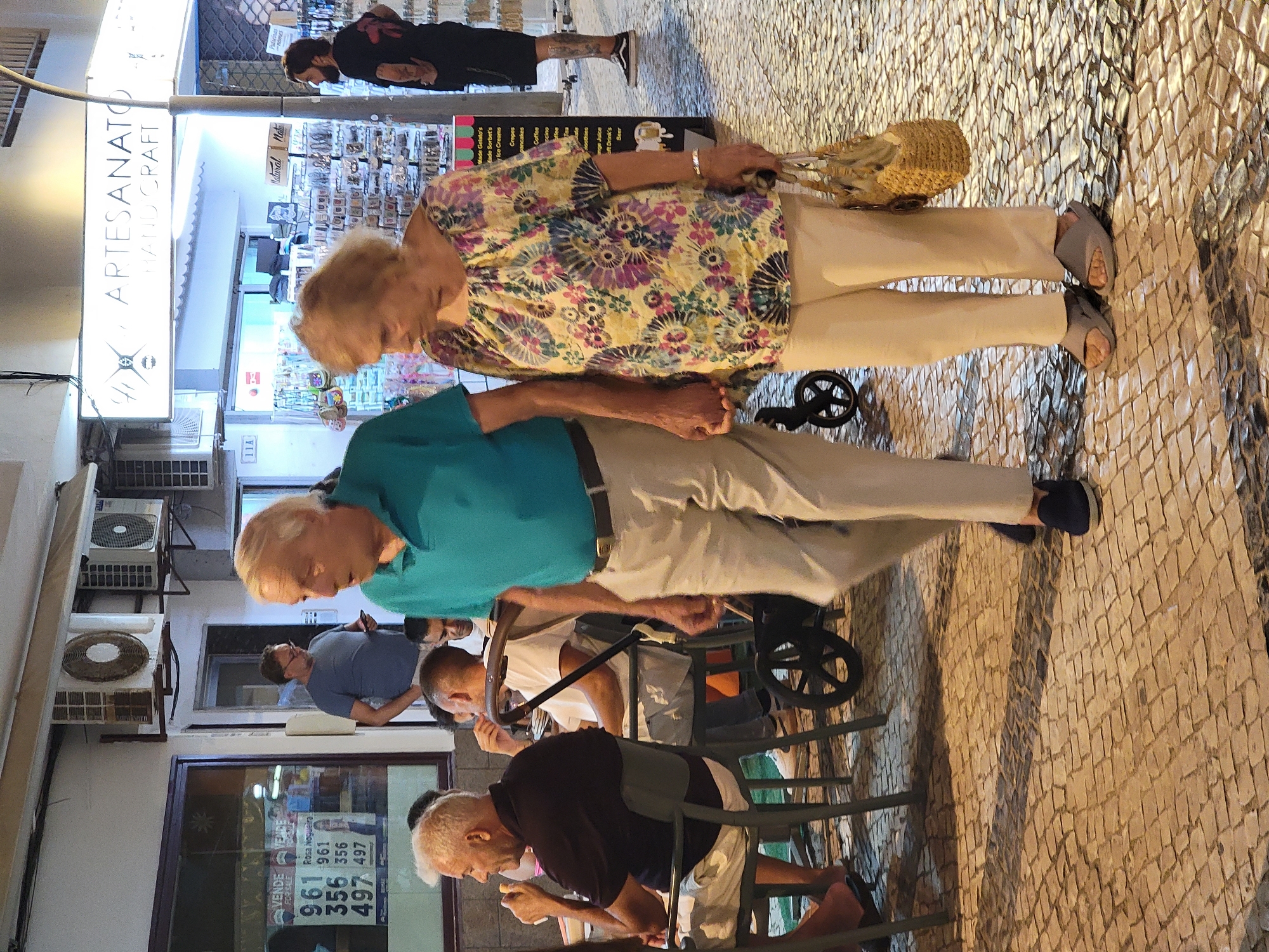 An elderly couple is walking hand-in-hand on a cobblestone street in Quarteira, Portugal. The man, on the left, is wearing a blue polo shirt and light-colored pants, with dark blue loafers. The woman, on the right, is wearing a brightly colored floral print blouse and light-colored pants with gray slip-on sandals. She is carrying a woven straw handbag. Behind them, on the right, a man with dark hair and a beard, wearing a black t-shirt and shorts, stands looking to the left. In the background, there is a storefront with "ARTESANATO HAND CRAFT" signage above it, and further to the left, a window display with real estate listings. The scene appears to be an outdoor commercial area, possibly a pedestrian walkway lined with shops. The lighting suggests it is either evening or a very overcast day.