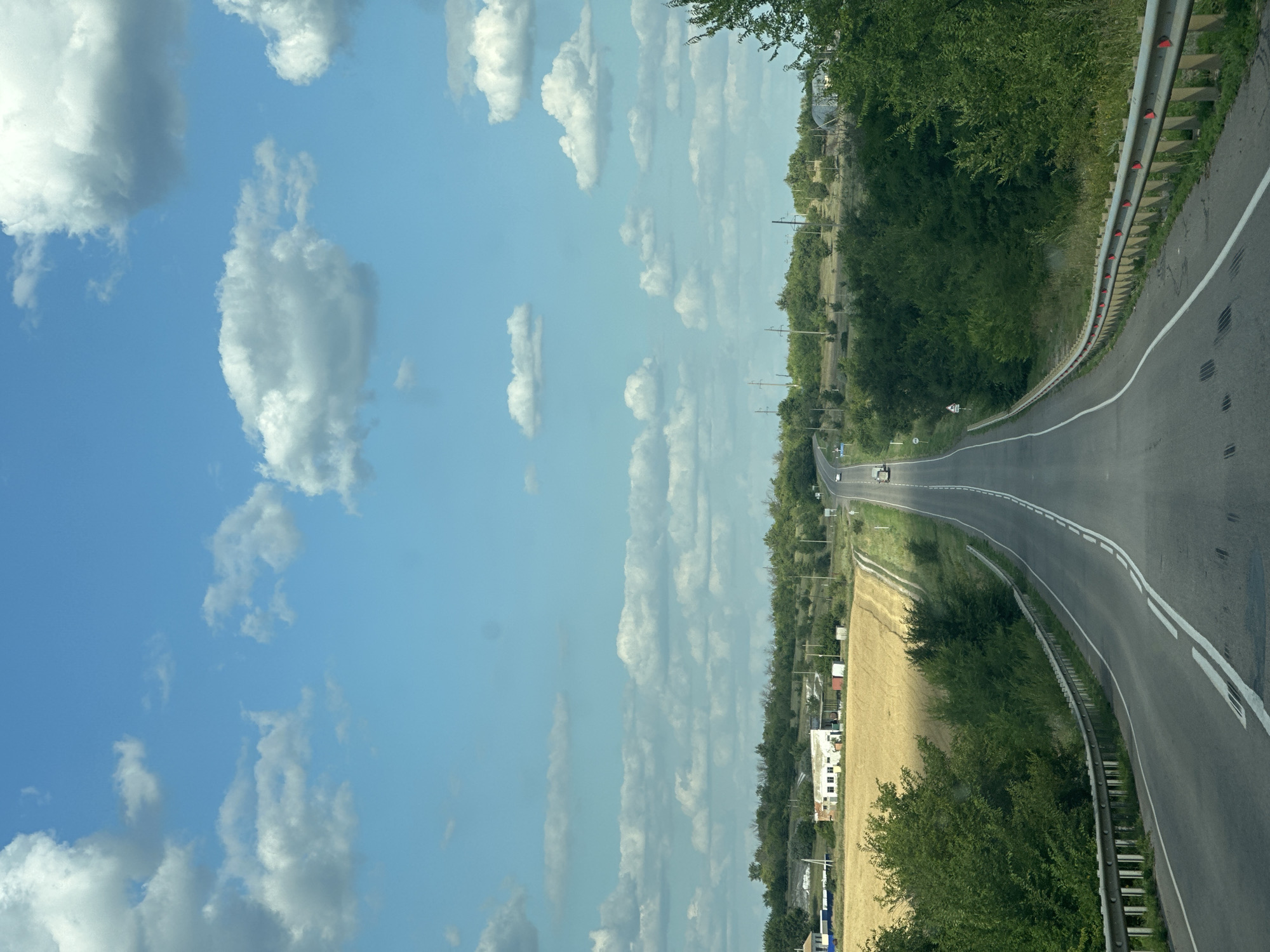 The image depicts a rural landscape with a paved road curving through rolling hills. The road is lined with a metal guardrail on the right side, and the surrounding vegetation consists of green trees and bushes, with a field of dry, golden crops to the left. In the distance, there are a few small buildings, possibly farm structures. The sky is a bright blue with scattered white cumulus clouds. Several vehicles are visible on the road, including cars and a truck. The overall scene suggests a sunny day in a countryside setting.