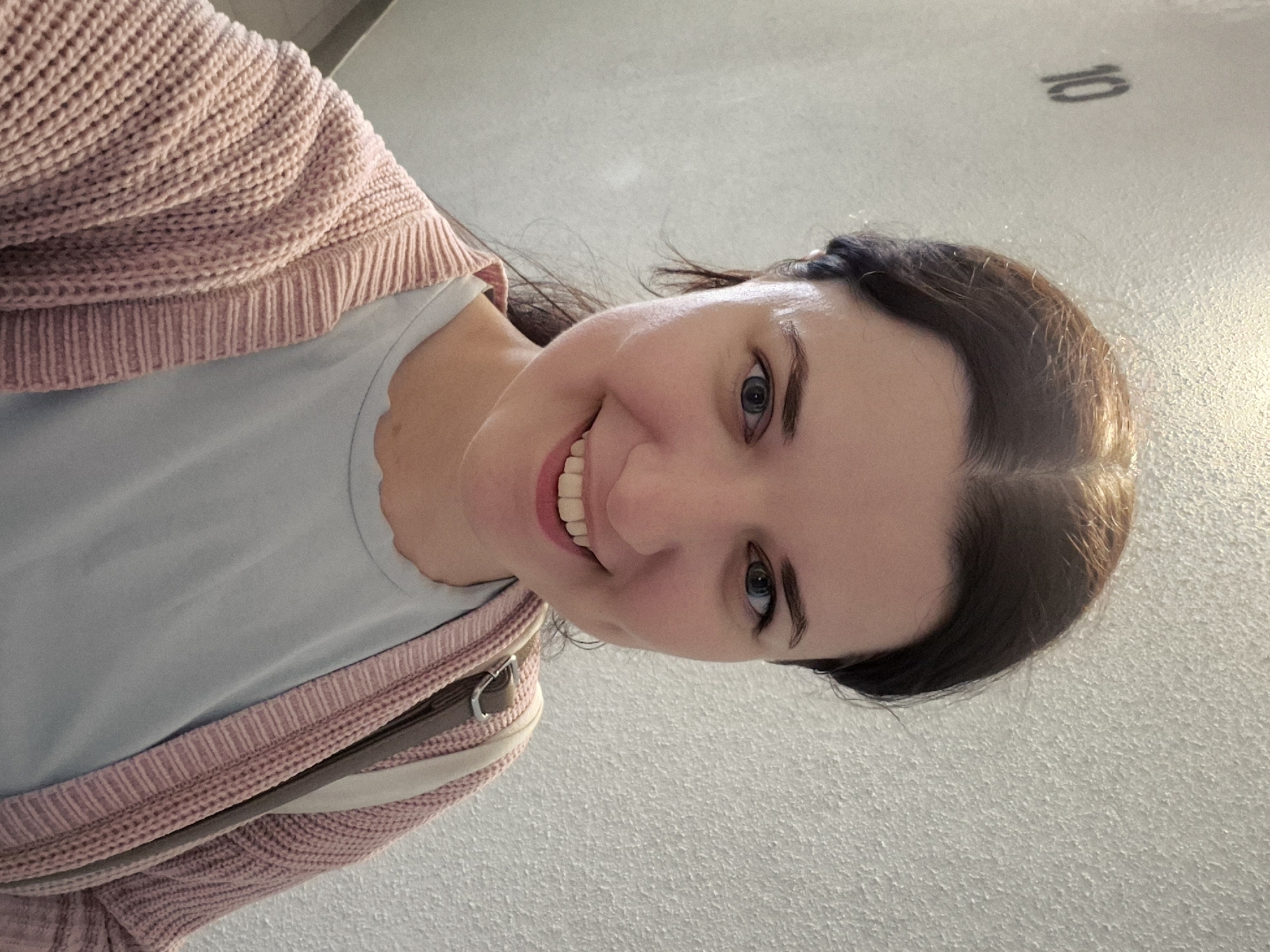 A woman with dark brown hair pulled back is smiling and looking at the camera in a selfie. She is wearing a light blue t-shirt and a pink knitted cardigan. A strap from a bag is visible over her right shoulder. The background is a textured wall with the number "10" stenciled on it. The lighting suggests an indoor setting.