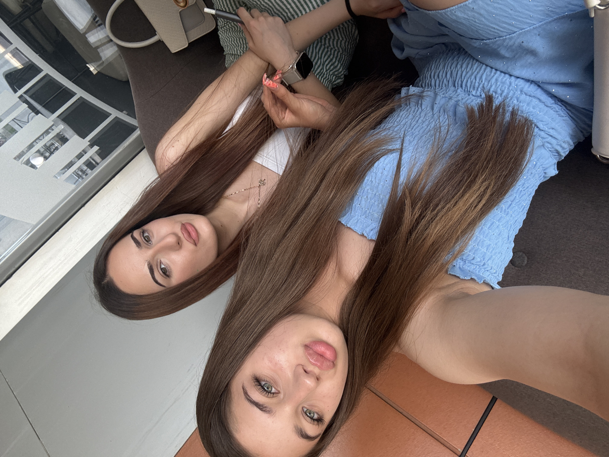 Two young women are featured in a close-up, horizontally oriented image, likely a selfie. Both women have long, straight brown hair.
The woman in the foreground has fair skin, green eyes, and full, light pink lips. She is looking directly towards the camera with a neutral expression. She wears a light blue, possibly strapless or spaghetti-strap top, which features a shirred or ruched texture around the bust and midriff, and appears to have subtle sparkling details. Her arm is visible in the lower right corner, resting on a brown tiled surface.
The woman in the background lies beside her, with her head positioned further up and to the left. She has brown eyes and is looking towards the camera with a relaxed expression. She wears a simple white top and a delicate gold-toned necklace with a small charm. Her left hand is visible, wearing a silver-toned smartwatch with a dark band. She holds a slender, dark object, possibly a vape pen or stylus, in her right hand. Her nails are painted with a vibrant, possibly patterned, design in orange/pink tones. She is partially covered by a striped green and white garment or cushion, and a light beige handbag with a gold-toned clasp rests near her.
The setting appears to be indoors, likely a cafe, lounge, or private residence. A dark upholstered sofa or seating area is visible behind them. To the left, there's a window or glass partition with a white frame and frosted or patterned elements, suggesting an indoor environment with natural light. The women appear to be relaxing and posing for the photo. No visible text is present in the image.