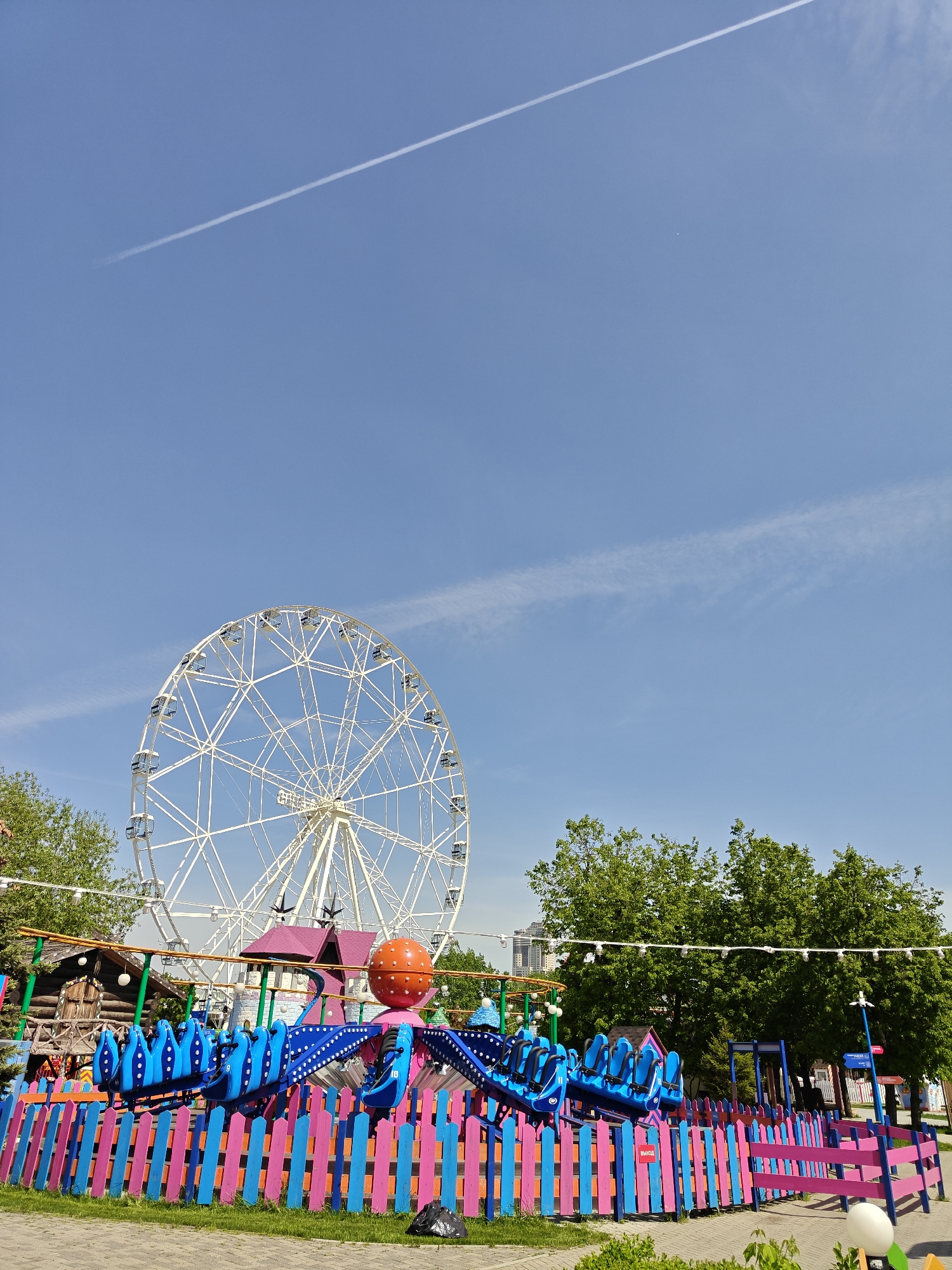 The image depicts a bright and sunny day at a park in Khimki, Russia, with a Ferris wheel as a prominent background feature. In the foreground, there is a children's amusement ride, enclosed by a colorful fence of blue, pink, and orange vertical slats. The ride appears to be a spinning or swinging type, with blue seats. A few small wooden structures, possibly related to the park or ride theming, can be seen behind the ride. Trees with green foliage frame the scene on both sides, and a contrail is visible in the clear blue sky.