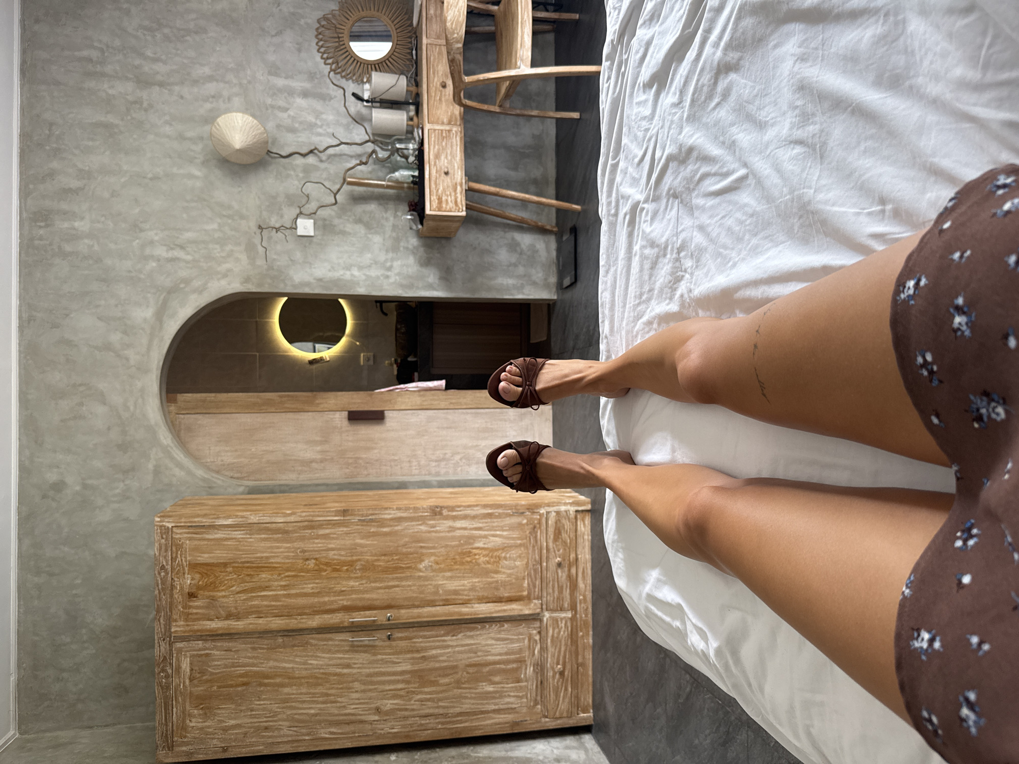 The image shows a person's legs and feet clad in brown sandals lying on a white bed inside a room in Tibubeneng, Indonesia. The room has gray textured walls and features a wooden dresser, a lighted mirror recessed into an arched alcove, and a rustic decorative arrangement consisting of a straw hat, a circular woven mirror, and a wooden structure.