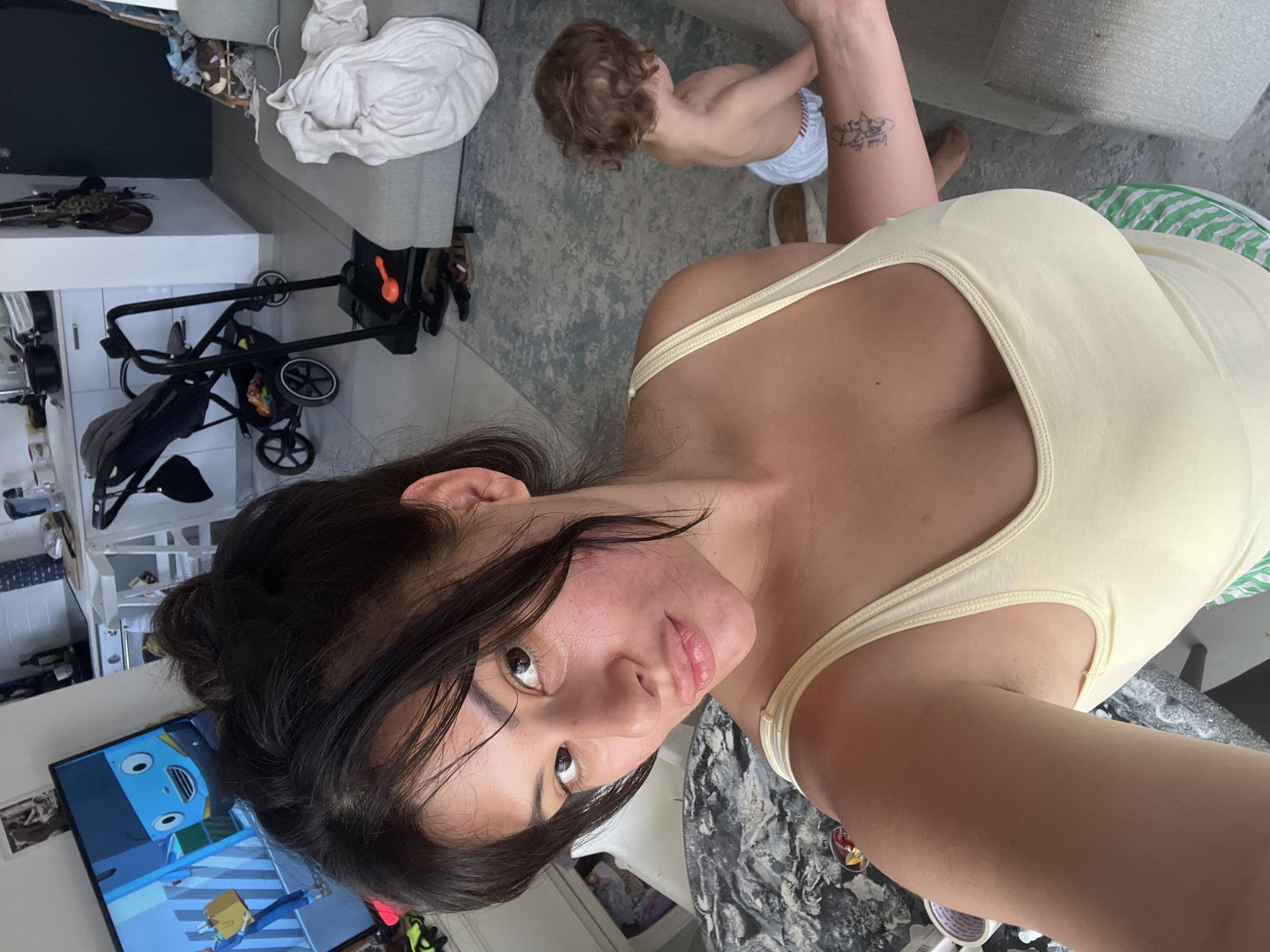 In this indoor shot, a woman and a young child are the primary subjects. The woman is in the foreground, taking a selfie. She has dark hair and is wearing a light yellow tank top and striped shorts. A tattoo is visible on her arm. The child, a toddler, is in the background, wearing a diaper and facing away from the camera.
The scene appears to be a living area, with a couch and a rug on the floor. A stroller is visible on the left side of the image, along with some household items and a television screen displaying a cartoon. The lighting suggests it is daytime. The overall mood seems casual and domestic. No text is clearly visible in the image. The location context is Kiryat Bialik, Israel.