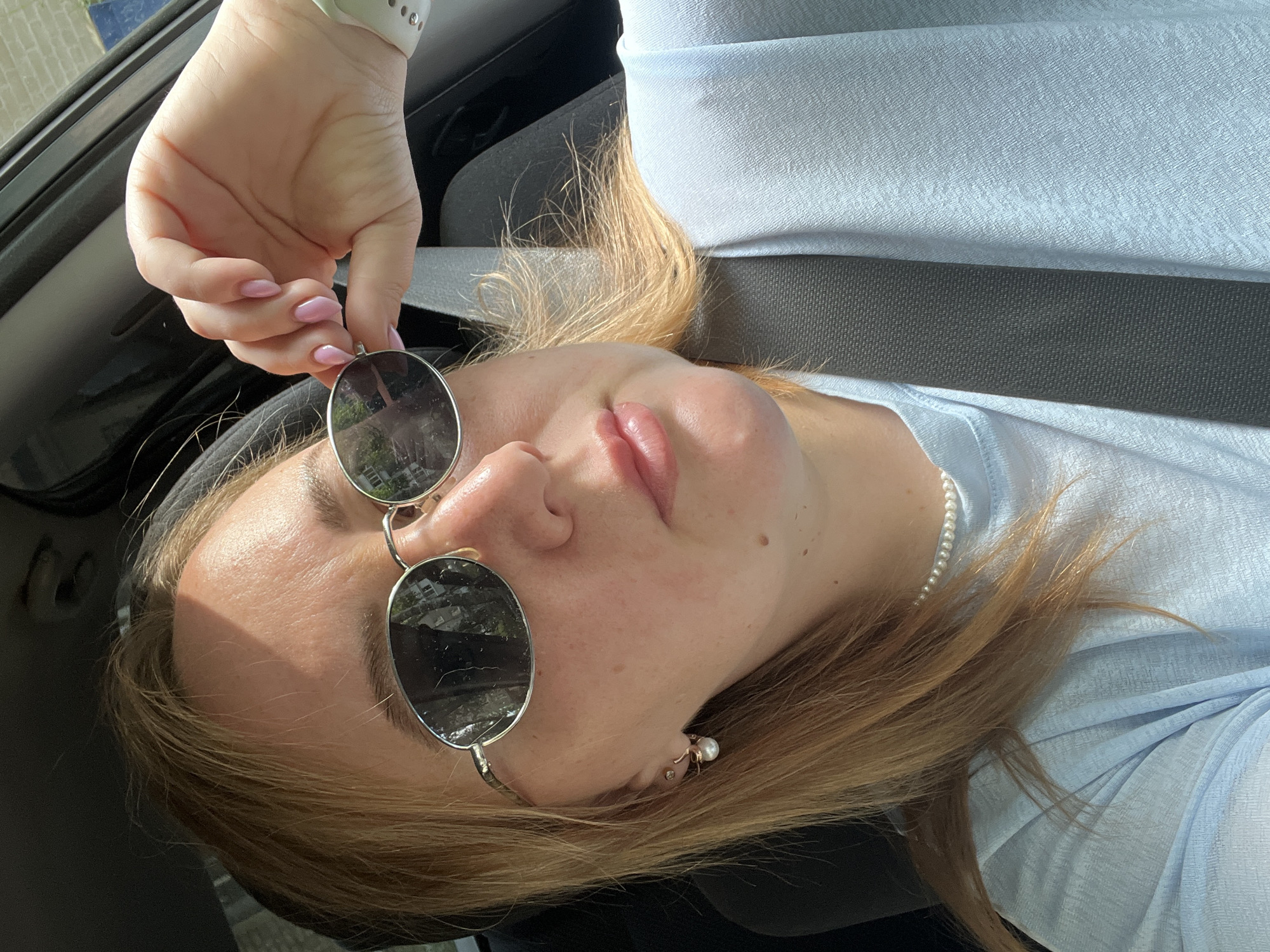 This is a close-up, overhead shot of a young woman reclining in what appears to be a car. She is wearing a light blue, flowy top, and a pearl necklace. She has blonde hair and is wearing sunglasses, which are being adjusted by her hand. Her fingernails are painted a pale pink. There is a seatbelt visible across her chest. The lighting suggests it is daytime, likely sunny, casting shadows on her face. The woman's expression is relaxed, with her eyes closed and lips slightly parted. There are no visible text elements in the image.