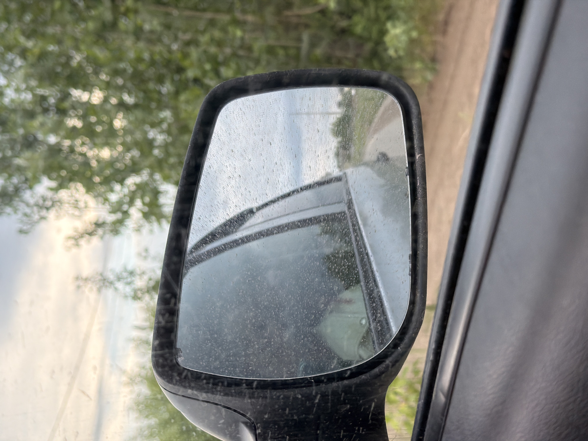 A close-up photograph taken from inside a vehicle shows a dusty side mirror reflecting a car behind it on a rural road. The road appears unpaved and is bordered by trees. The sky is overcast. The vehicle's interior, specifically the door panel, is visible to the right of the mirror. Given the requirement for the location being Chaikovsky, Russia, the photograph could depict a rural road near the city.