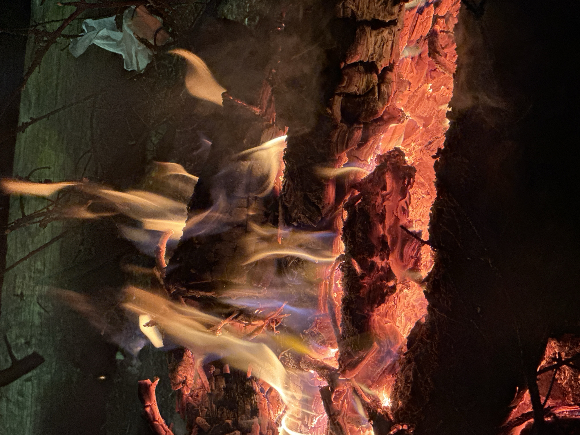 The image depicts a close-up of burning wood. The wood is predominantly reddish-orange in color, with deep cracks and grooves. Flames and smoke are visible, emanating from the wood. Some parts of the wood are charred black. The background is largely dark and indistinct. A pale green wooden object, possibly a fence post, appears on the left side of the image, with branches and a small piece of white cloth hanging from it. The location of Юзуфоўскі сельскі Савет, Belarus, cannot be confirmed. There are no people, interactions, or visible actions other than burning, and no discernible subjects or objects other than wood and fire.