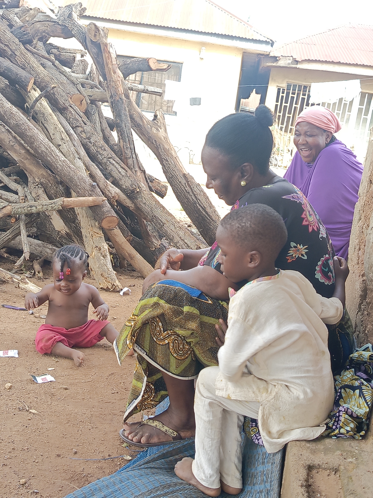 Outside a mud structure in Akwanga, Nigeria, a pile of logs rests to the left of a seated woman in a black and floral patterned dress and sandals. She is interacting with a standing boy wearing a white tunic and pants. A woman with a pink head covering wearing a purple garment sits to the right of the first woman. A baby in red shorts sits on the ground, near paper debris. Behind them is a building with a corrugated metal roof and barred window.