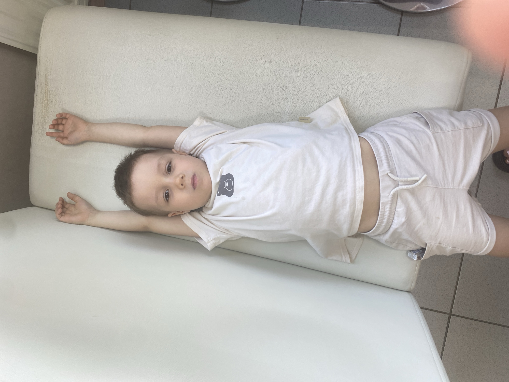 A young boy is lying on a white padded surface, possibly a bench or a padded section of a chair. He is wearing a light-colored t-shirt with a graphic on it and matching shorts with a drawstring. He is lying on his back with his arms stretched out to the sides. The floor is tiled. The image appears to have been taken indoors in the city of Chaikovsky, Russia.