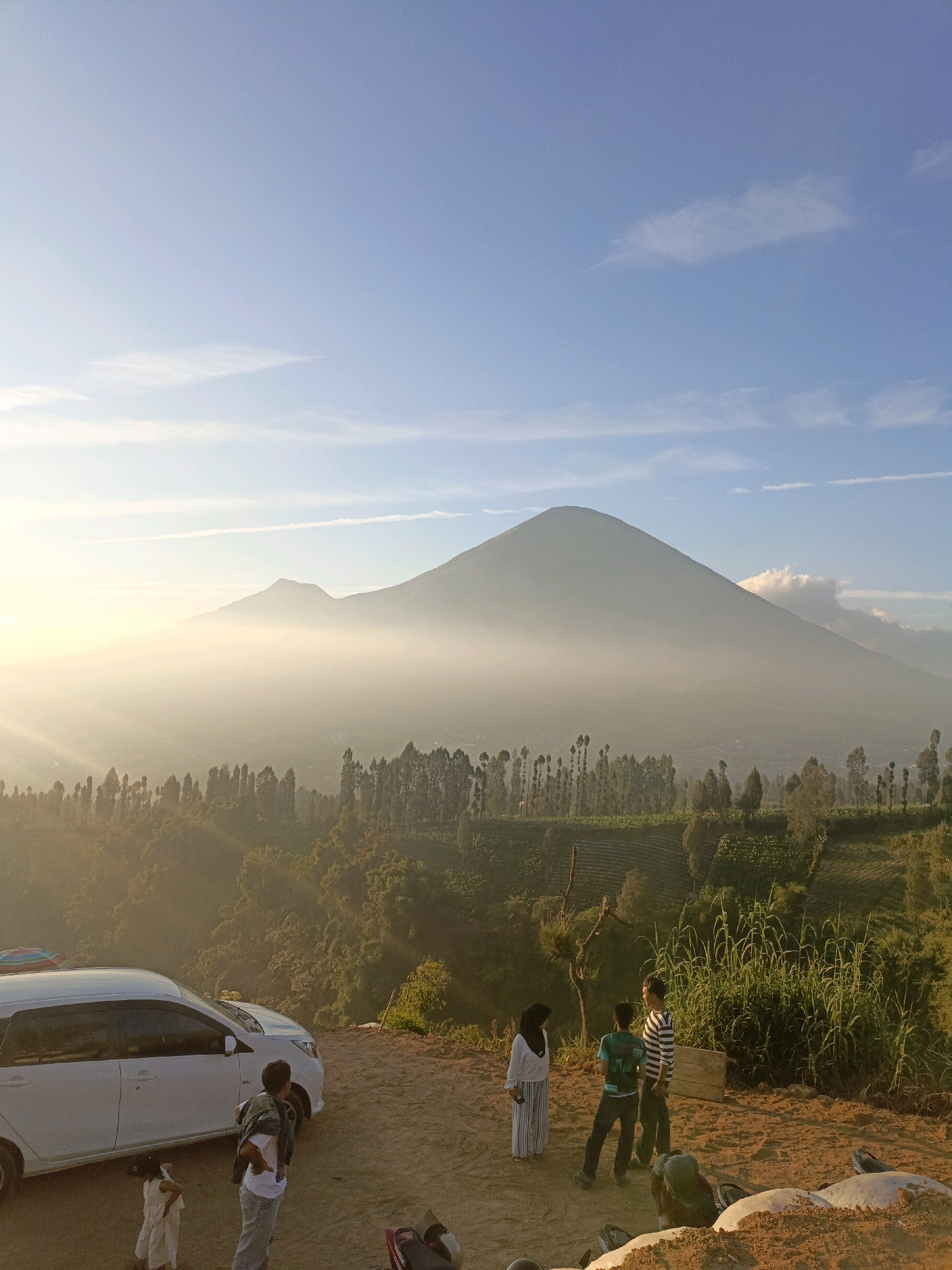 The image depicts a scenic view of Mount Sumbing in Butuh Kidul, Indonesia, at sunrise or sunset. The sky is a clear blue with some wispy clouds. In the foreground, a white car is parked on a dirt road, with several people standing near it. A woman in a white hijab and striped dress stands to the left, looking towards the mountains. Two young men are talking near the car, one wearing a green and blue striped shirt and the other a black and white striped shirt. The landscape is characterized by lush green hills and terraced fields, with a dense forest of tall, slender trees in the middle ground. The distant mountains are partially shrouded in a soft haze, with a hint of sunlight breaking through on the left side of the frame. The overall atmosphere is tranquil and picturesque, capturing the natural beauty of the Indonesian countryside.