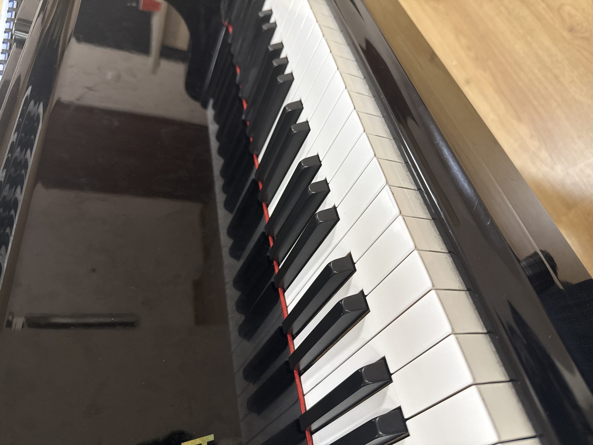 The image shows a close-up, high-angle perspective of piano keys. The keys are predominantly white with black accidentals. The piano appears to have a glossy, black finish, with reflections visible on its surface. The background includes a blurry, neutral-toned wall or floor to the right and a reflection of a room on the left side. No people are visible. The setting does not provide information about the city of Sofia, Bulgaria.