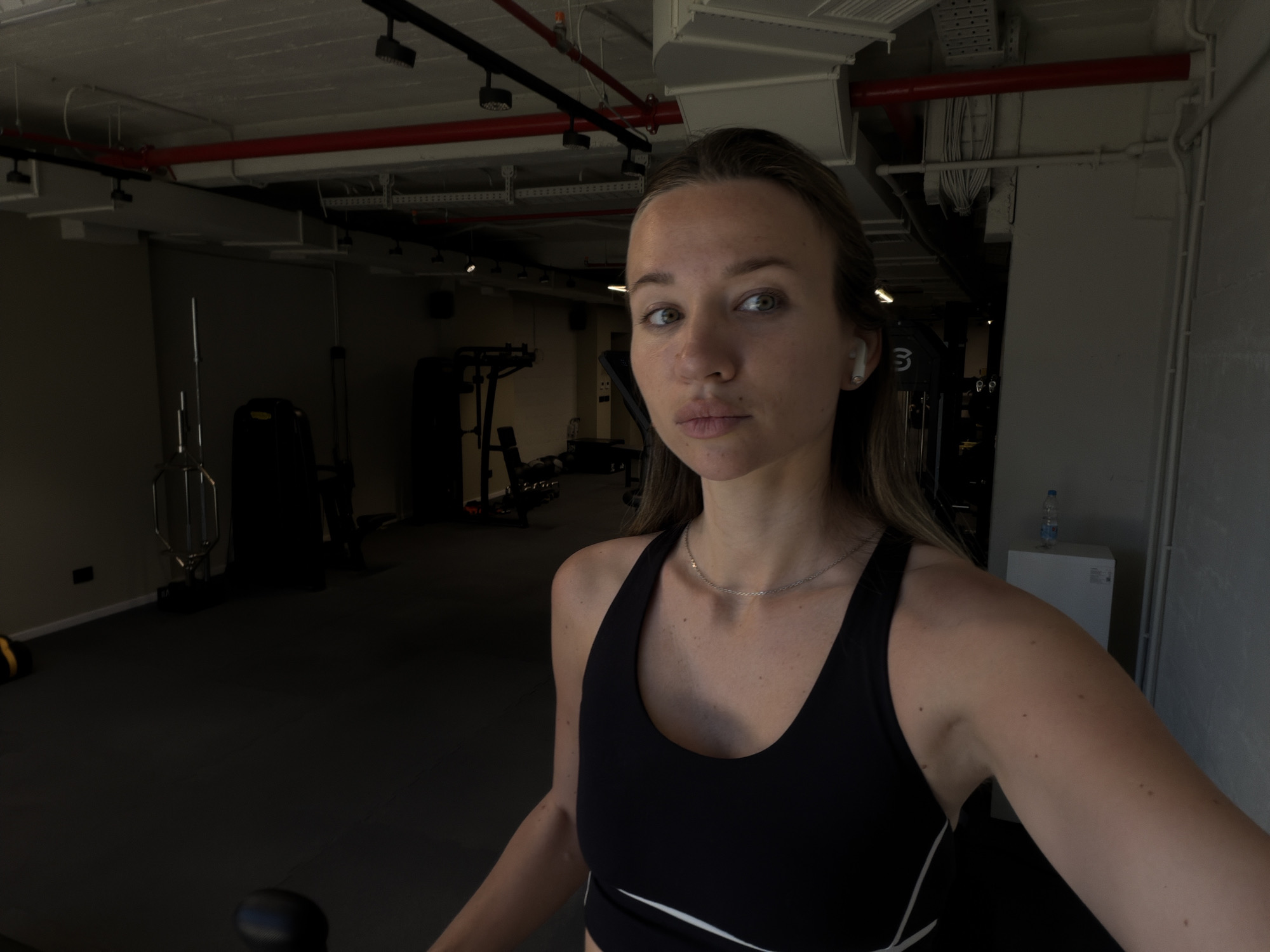 A medium-shot captures a woman in a fitness setting. She is fair-skinned, with shoulder-length brown hair and is wearing a black sports bra with a white accent line. An earbud is visible in her ear.
The background indicates the location is a gym or fitness center. Various exercise machines and equipment are visible, including a weight machine. The gym features a dark floor and lighter-colored walls, with exposed ceiling structures.