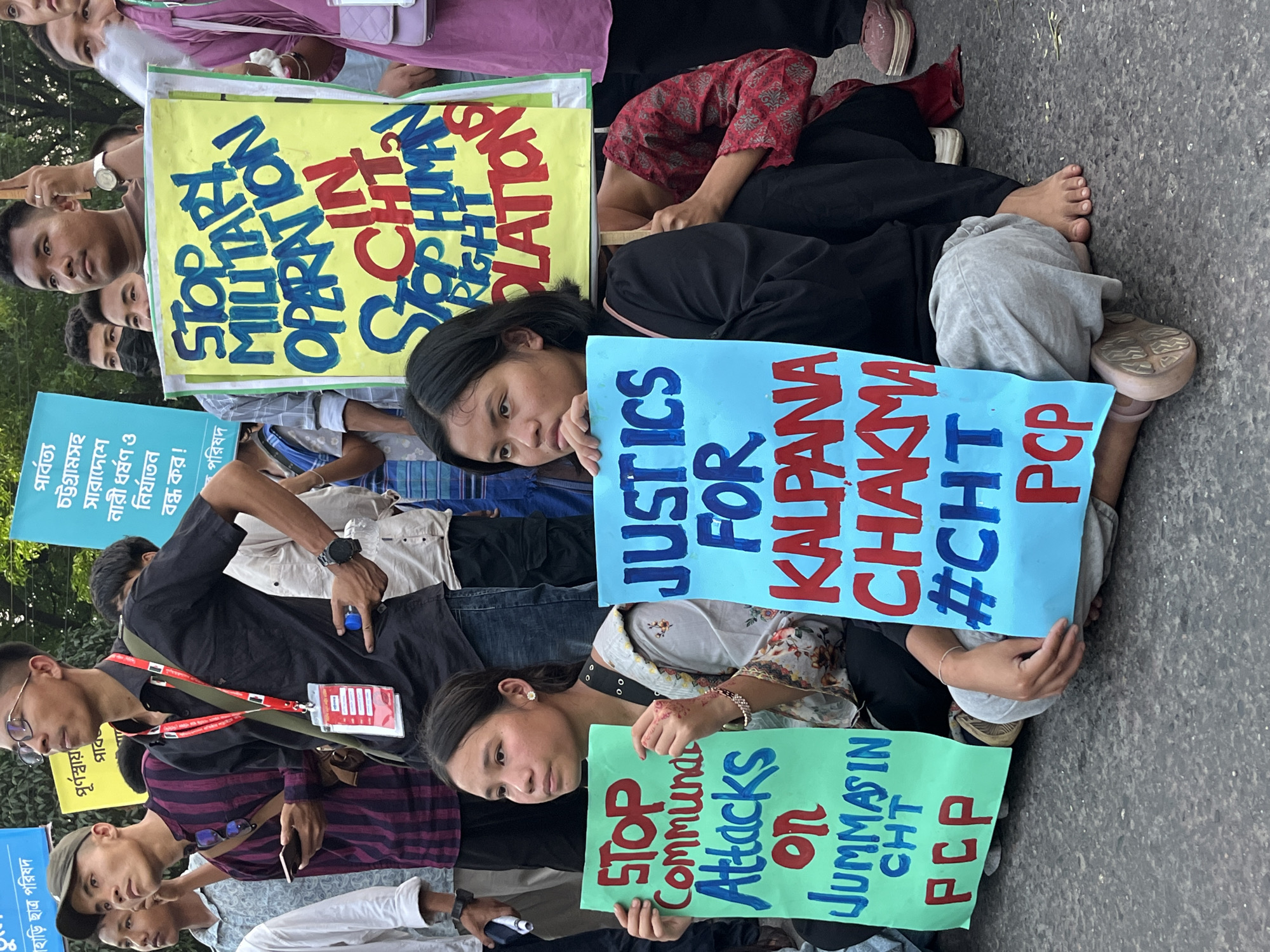 A daytime street scene in Dhaka, Bangladesh, shows a group of people protesting. They are seated on the ground, holding hand-painted signs. The signs bear messages in English, demanding an end to military operations, an end to communal attacks, and seeking justice for Kalpana Chakma. The signs include phrases like "Stop Military Operation CHT Stop Human Right Violation," "Stop Communal Attacks on Jummasin CHT PCP," and "Justice for Kalpana Chakma #CHT PCP." The protesters appear to be a mix of young men and women. One individual is wearing a lanyard with an identification card. The setting is an urban environment, evidenced by the asphalt road.
