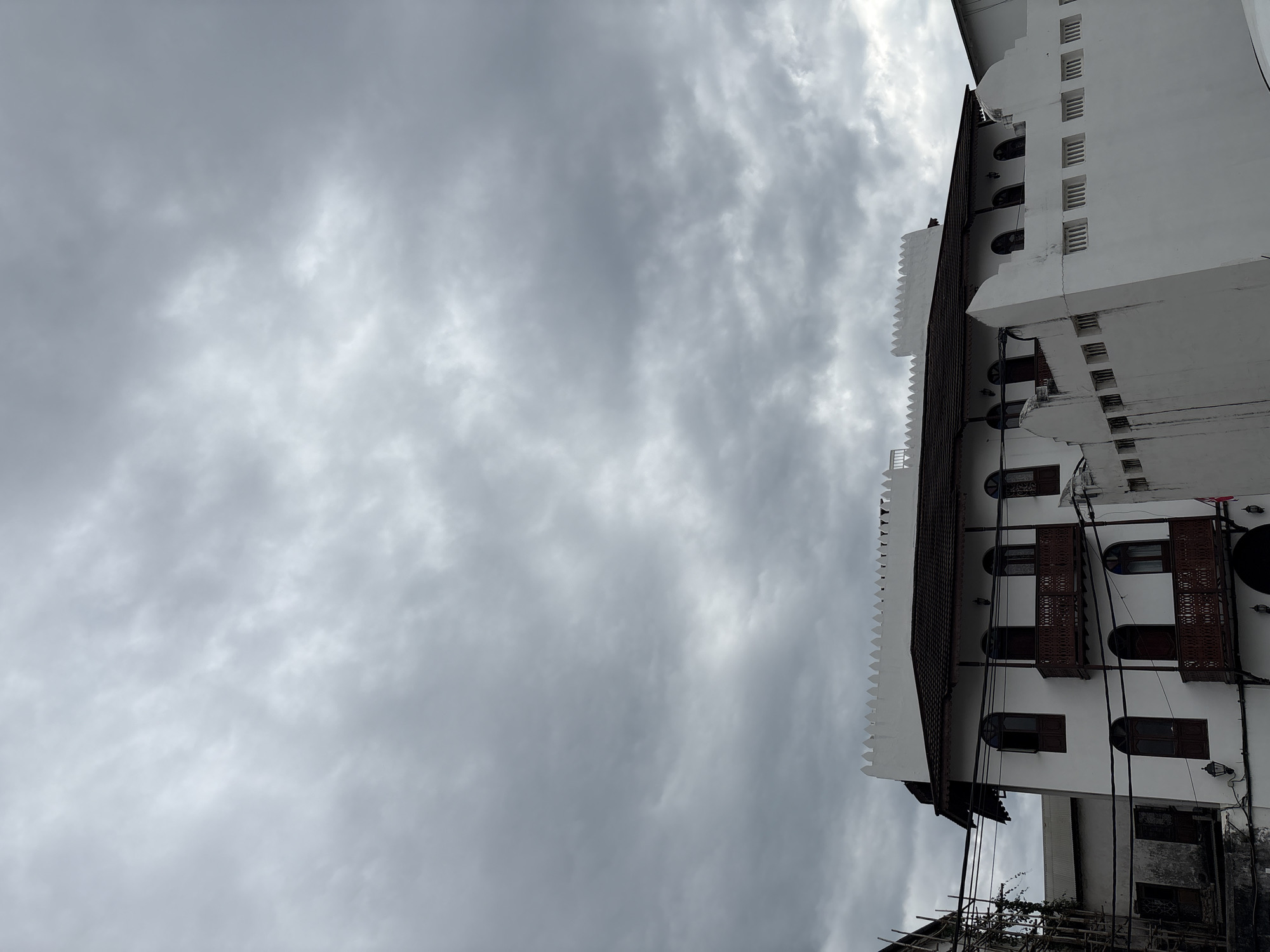 In Kelele Square, Tanzania, a three-story white building with arched windows and wooden balconies stands under a cloudy sky. The building has a dark tiled roof and a decorative crenellated parapet. To the left of the building, scaffolding is visible, suggesting ongoing construction or renovation. A lower white wall with small, rectangular openings runs in front of the main building. Electrical wires are strung across the scene. The sky is filled with heavy, gray clouds.