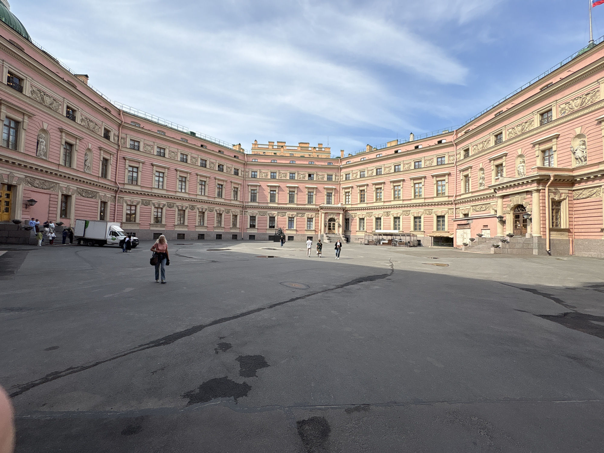 The image depicts a large, open courtyard surrounded by an ornate, multi-story building in Saint Petersburg, Russia. The building has a distinctive pink facade with numerous windows, decorative moldings, and sculptural elements. The courtyard is paved with asphalt, showing signs of wear and repair. Several people are present, including individuals walking across the courtyard and a small group near a white truck parked on the left side of the frame. The sky is partly cloudy, suggesting daylight conditions. A flag is visible on the right side of the image.