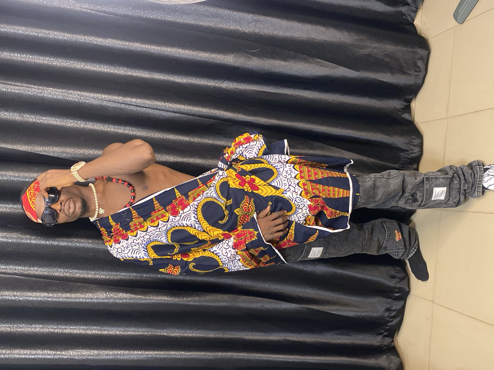 The image shows a full-body shot of a man standing in front of a black curtain. He is wearing a red bandana, sunglasses, and a pearl necklace with a red and black beaded necklace over it. He has a colorful, patterned fabric draped over his left shoulder and across his torso. He is wearing dark wash jeans with unique detailing and black socks. The man is adjusting his bandana with his right hand. The background is a dark, textured curtain, and the floor is tiled. The overall lighting suggests an indoor setting. The man's attire and pose convey a sense of style and cultural expression. There is no identifiable information within the image that specifies the location as Abakaliki, Nigeria.