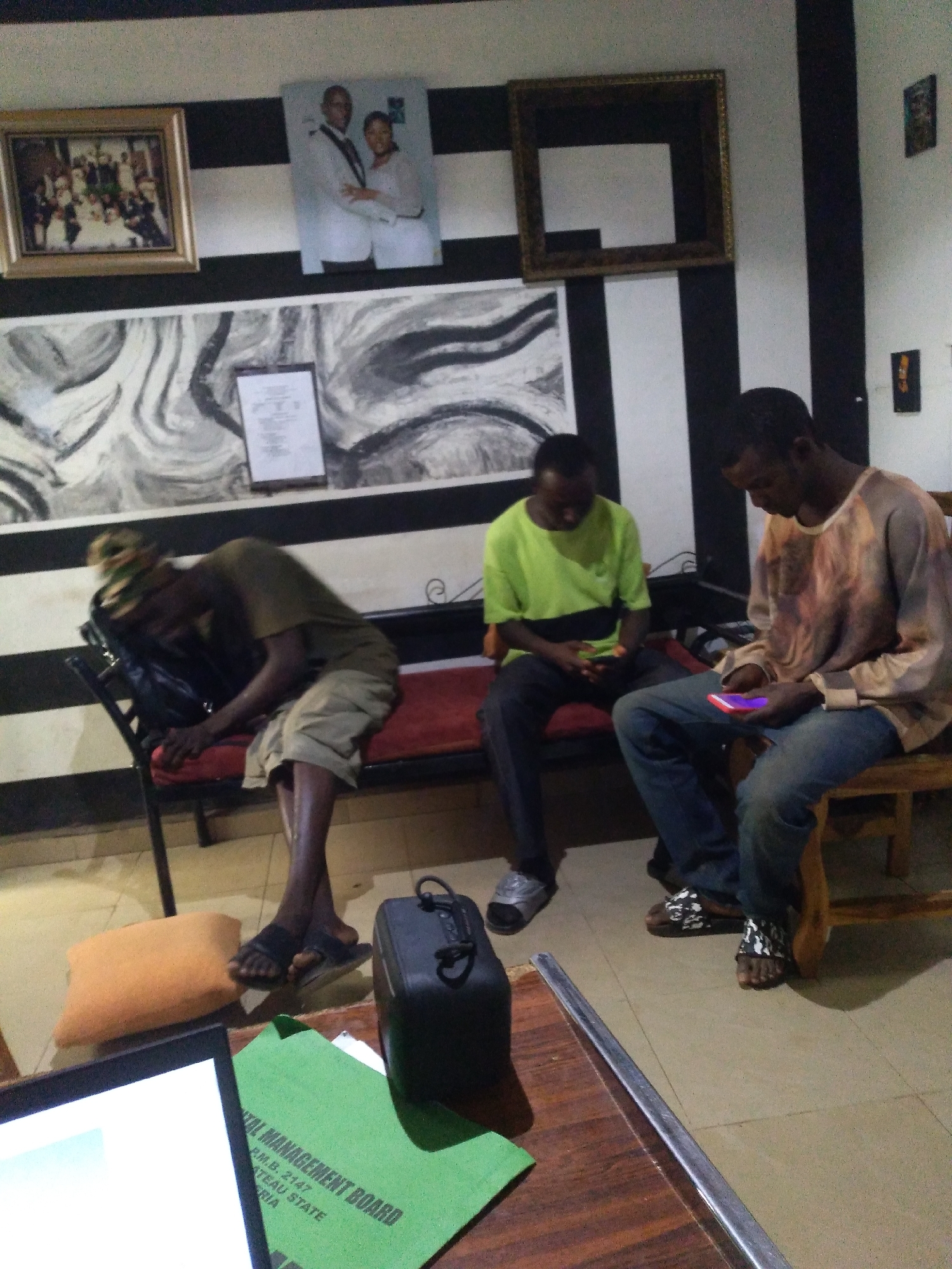 The image depicts an indoor scene, likely in a waiting area or common space, with a strong emphasis on black and white striped walls. The room is sparsely furnished, featuring a long, red-cushioned bench. On the bench, three individuals are present.
On the left, a person is slumped over, seemingly asleep or resting, with a dark bag next to them. In the center, another person is seated, looking down at their phone. To the right, a third individual is also seated, looking at their phone, which emits a purple glow.
The walls are adorned with several framed pictures and a large abstract artwork. A framed photograph of a couple stands out, as does a gold-toned empty frame. A document is also visible, attached to the wall. In the foreground, a wooden table has a laptop and a green folder with text, including "MENT BOARD" and "PLATEAU STATE."
The lighting appears to be artificial, casting shadows that contribute to the overall atmosphere of the scene. The presence of a laptop and phones suggests a modern setting, contrasting with some of the decorative elements. The details about "PLATEAU STATE" on the folder strongly suggest the location of Jos, Nigeria.