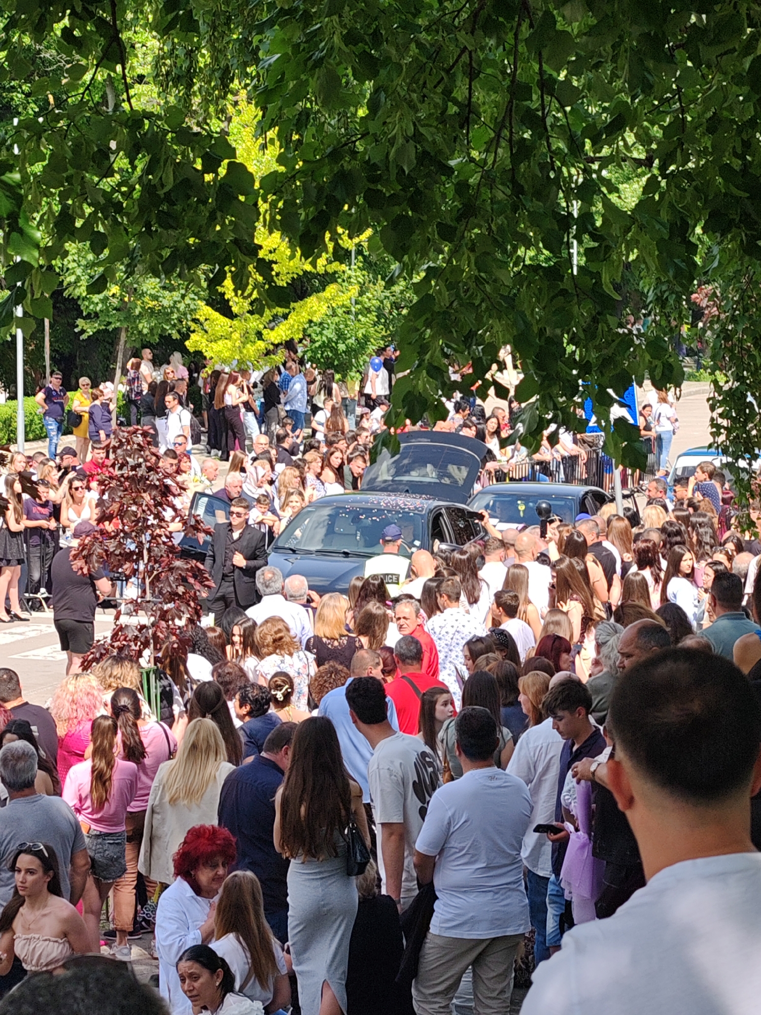 The image shows a large crowd of people gathered on a street in Shumen, Bulgaria. The crowd surrounds a black car. People are wearing casual clothing, with a few individuals dressed in suits. A uniformed police officer is near the vehicle. The environment includes trees, with some branches hanging into the frame. The crowd's density suggests a significant event or gathering. The location, based on common knowledge, is Shumen, Bulgaria.