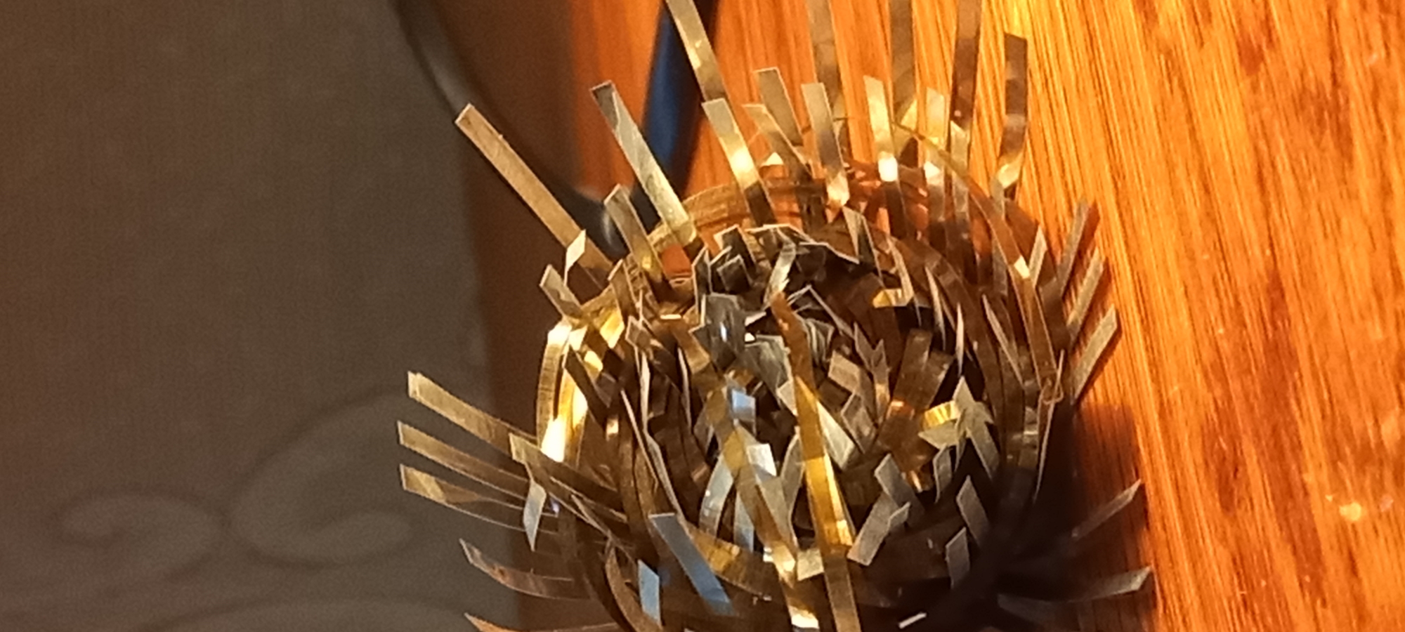 The image displays a close-up of an abstract, spherical object, likely a decorative item, set against an indoor backdrop. The central subject is a densely tangled mass of numerous narrow, reflective strips. These strips appear metallic, possibly Mylar or a similar material, with a dominant golden or brass-like hue, shimmering brightly due to strong reflections. They are intricately interwoven, forming a generally spherical shape, with many individual strips extending outwards, giving the object a spiky or bristly texture.
The object is positioned on a warm, reddish-brown wooden surface, which exhibits distinct wood grain patterns, suggesting it could be a table, shelf, or floor. In the background, there is a darker, indistinct area, likely a wall or another piece of furniture, providing a subtle contrast to the illuminated object.
No people are present, and no specific activity or event is depicted; the image is a still-life. The lighting suggests an indoor setting, possibly under artificial light, given the pronounced reflections on the metallic strips. There is no visible text in the image. While the context of Kryvyi Rih, Ukraine, is provided, there are no discernible visual cues within the image to indicate this specific geographical location.