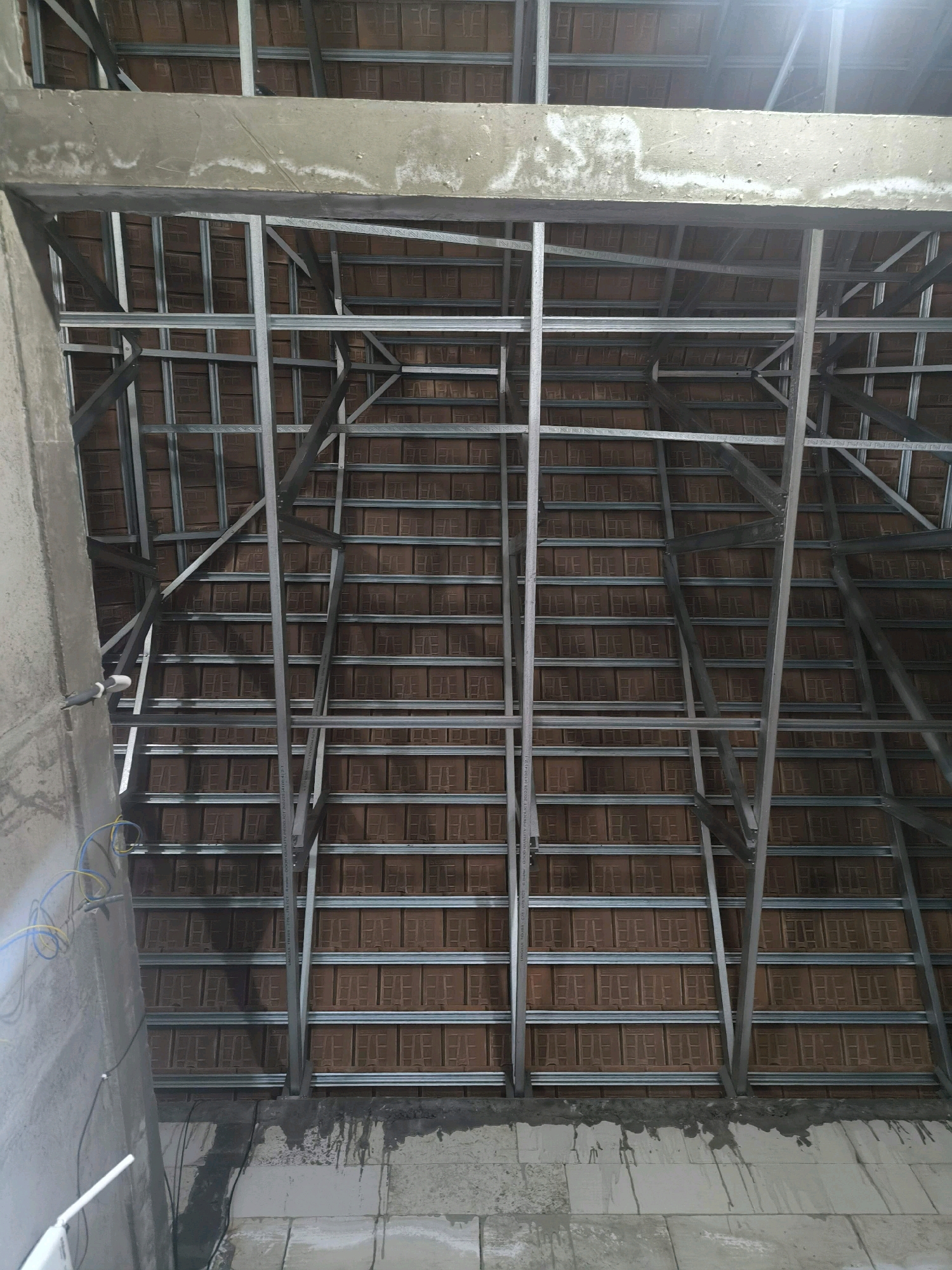 The image shows the underside of a roof structure under construction, likely in Tabanan, Indonesia. The roof features a metal framing system supporting terracotta tiles. A concrete beam runs horizontally across the top of the frame. Visible on the left is a concrete column, partially covered by gray material, with yellow and blue electrical wires extending from it. The lower section of the image shows a wall constructed from light-colored blocks, with dark streaks of what appears to be dried cement or water stains running down from the roof structure. The scene suggests an ongoing building or renovation project.