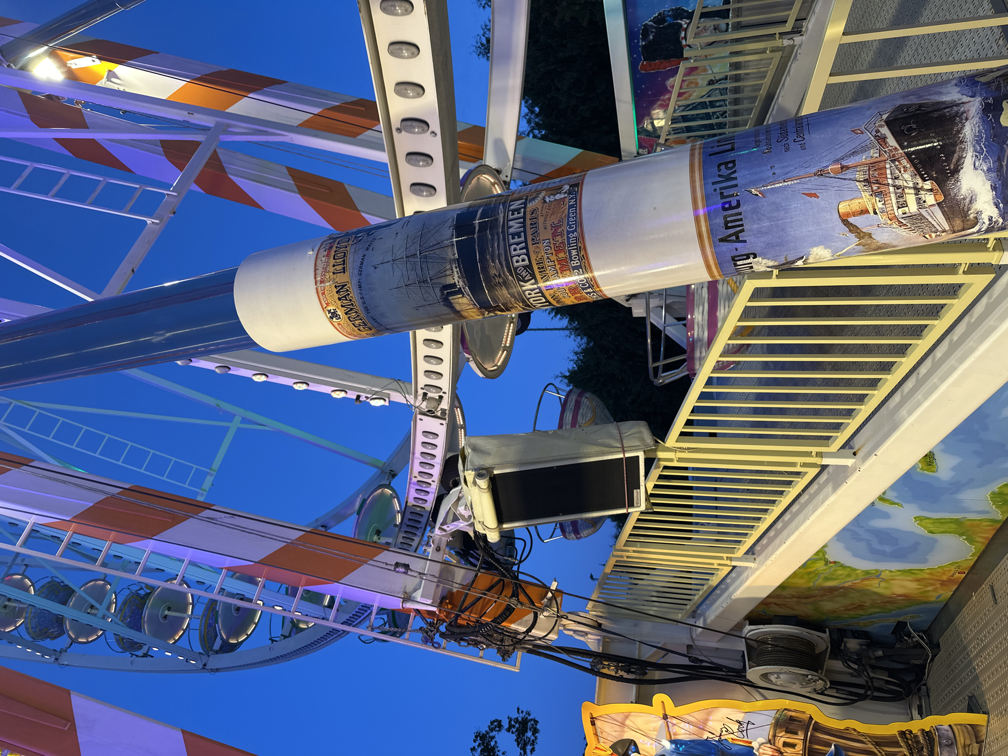 This image captures a close-up view of a carnival ride at the Brokser Erntedankfest in Bruchhausen-Vilsen, Germany. The ride features a large cylindrical structure wrapped with decorative imagery of historical ships and text related to shipping lines, specifically the "Norddeutscher Lloyd Bremen" and "Hamburg-Amerika Linie". The ride's structure, painted with orange and white stripes, is visible against a deep blue evening sky. In the background, parts of the Ferris wheel are discernible, along with other carnival attractions. A yellow railing lines a pathway leading up to the ride, and the base of the ride is adorned with a mural depicting a world map. The overall scene suggests a festive atmosphere at a fairground in the evening.