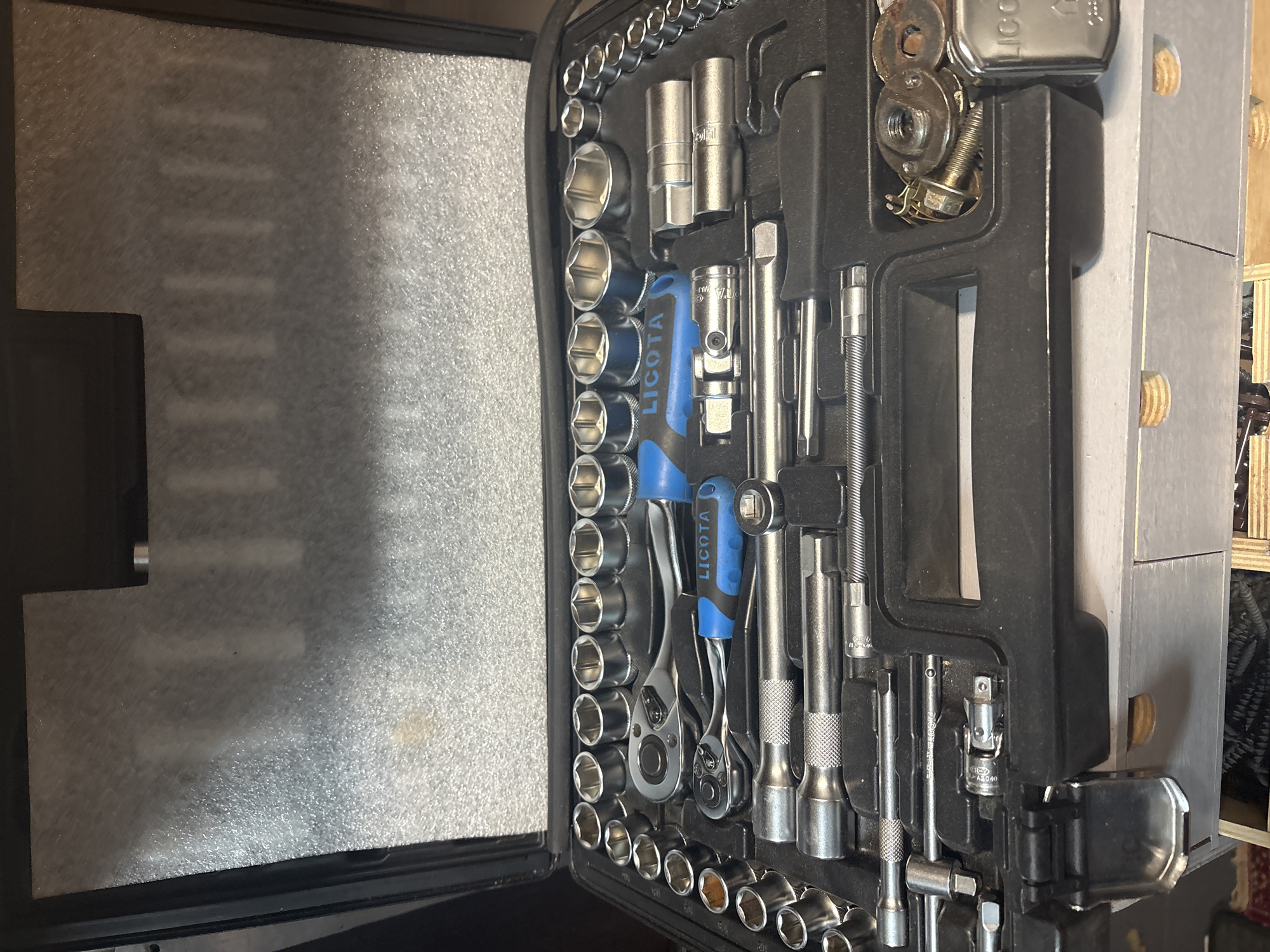 The image shows an open black toolbox filled with various metal tools, primarily a socket wrench set. The toolbox is resting on a light gray drawer unit, suggesting an indoor setting. The tools include a ratchet, extensions, and numerous sockets of varying sizes, many of them hexagonal. Some of the sockets are arranged in rows, while others are positioned within specific molded compartments in the black plastic interior of the toolbox. There are also a few other metal items, possibly nuts and bolts, in a compartment on the right side of the toolbox. The background inside the toolbox lid appears to be lined with a light-colored, textured foam material. The overall impression is of a neatly organized collection of mechanical tools. The image is taken from a slightly high angle, looking down into the toolbox. The location is identified as Березка Снт (поварово), Russia.