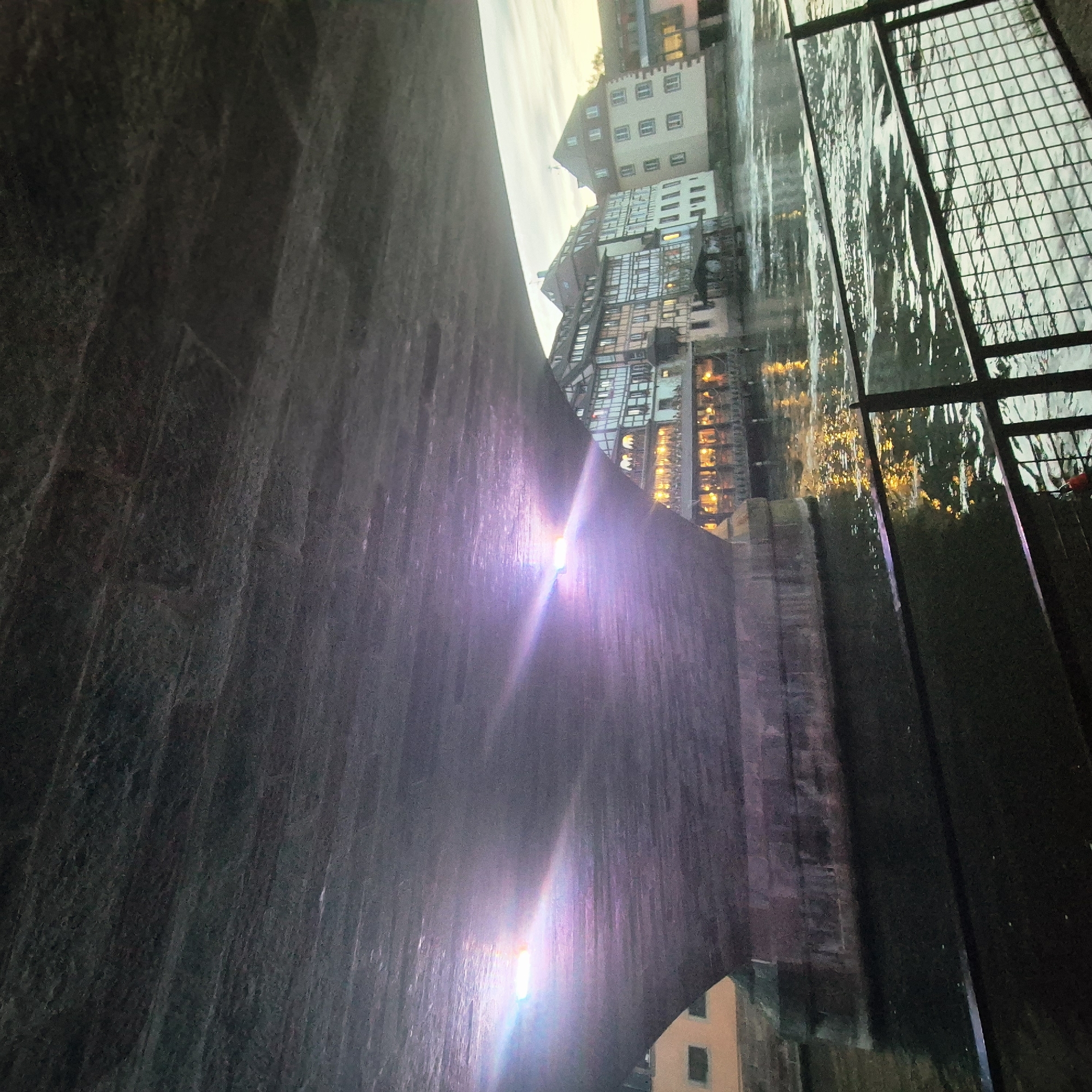 The image depicts a static scene viewed from directly beneath a large, dark, textured stone or concrete bridge spanning a river. This location is identified as the Petite France district of Strasbourg, France.
The underside of the bridge dominates the upper portion of the frame, displaying horizontal striations and varying shades of dark gray. Multiple bright, white and purple-hued light sources are visible on the bridge's underside, producing prominent lens flares that extend across the concrete surface.
Below the bridge structure, the Ill River flows with visible ripples and movement. On the immediate right, a dark, grid-patterned metal railing or fence extends vertically and horizontally, partially obscuring the water surface.
On the far bank, a series of multi-story buildings lines the river. These include characteristic Alsatian half-timbered houses with dark exposed beams, light-colored walls, and pitched roofs. One building features a brightly illuminated ground-floor or waterside extension, possibly a restaurant or cafe, with numerous warm yellow lights reflecting on the water. Another building appears lighter in color, possibly cream or white, with multiple uniformly spaced windows.
The sky, visible above the buildings, is a pale, overcast gray, suggesting twilight or early evening conditions. No people are visible within the frame, and no specific actions or interactions are discernible. The scene focuses on the architectural elements, the river, and the interplay of artificial light at dusk.