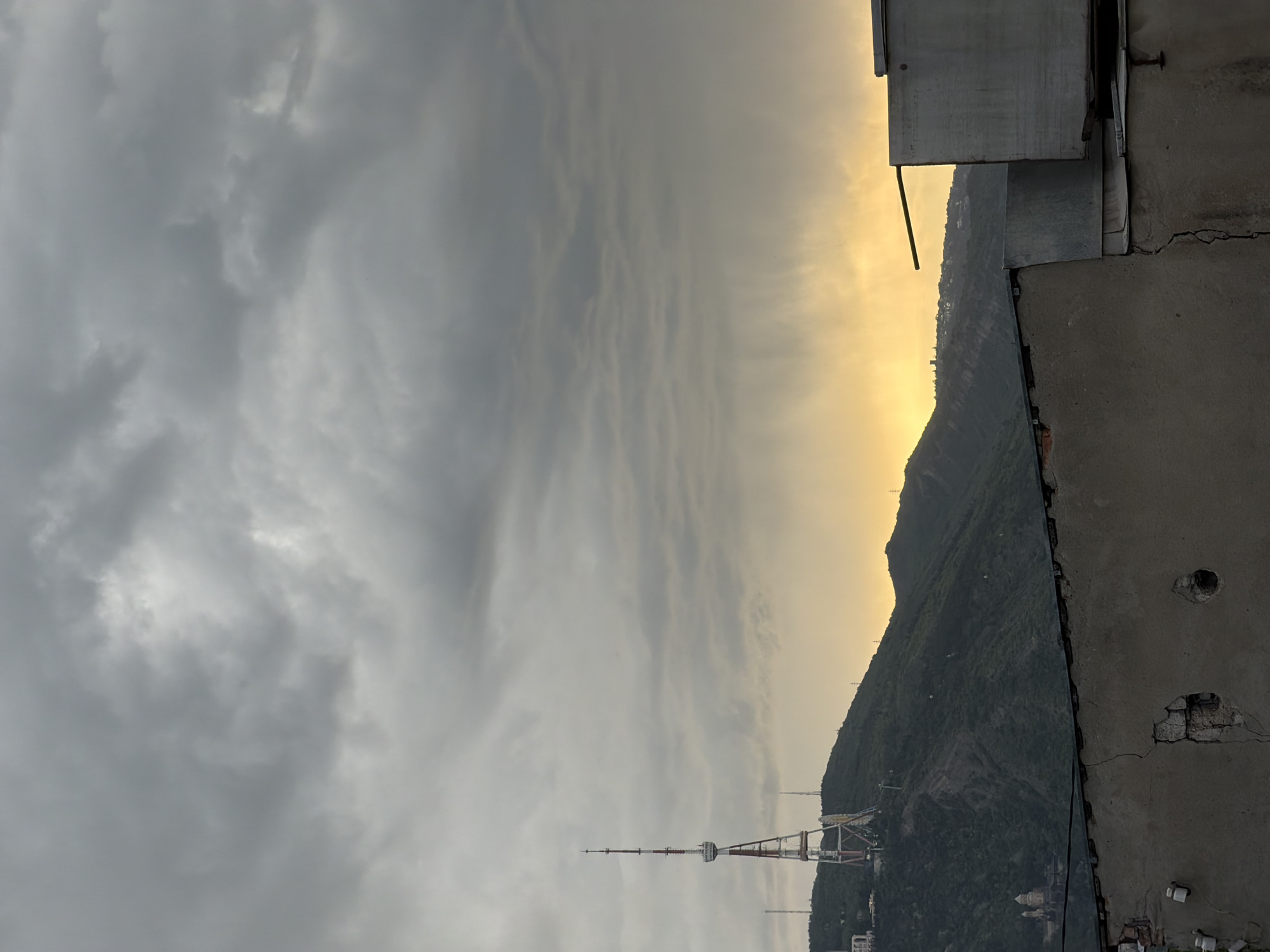 The image shows a view from a rooftop in Tbilisi, Georgia, looking towards the Tbilisi TV Broadcasting Tower. A mountain covered in green vegetation rises up in the background. The sky is cloudy and grey with a yellow glow where the sun is setting behind the mountain. A concrete rooftop and a metal structure are in the foreground.