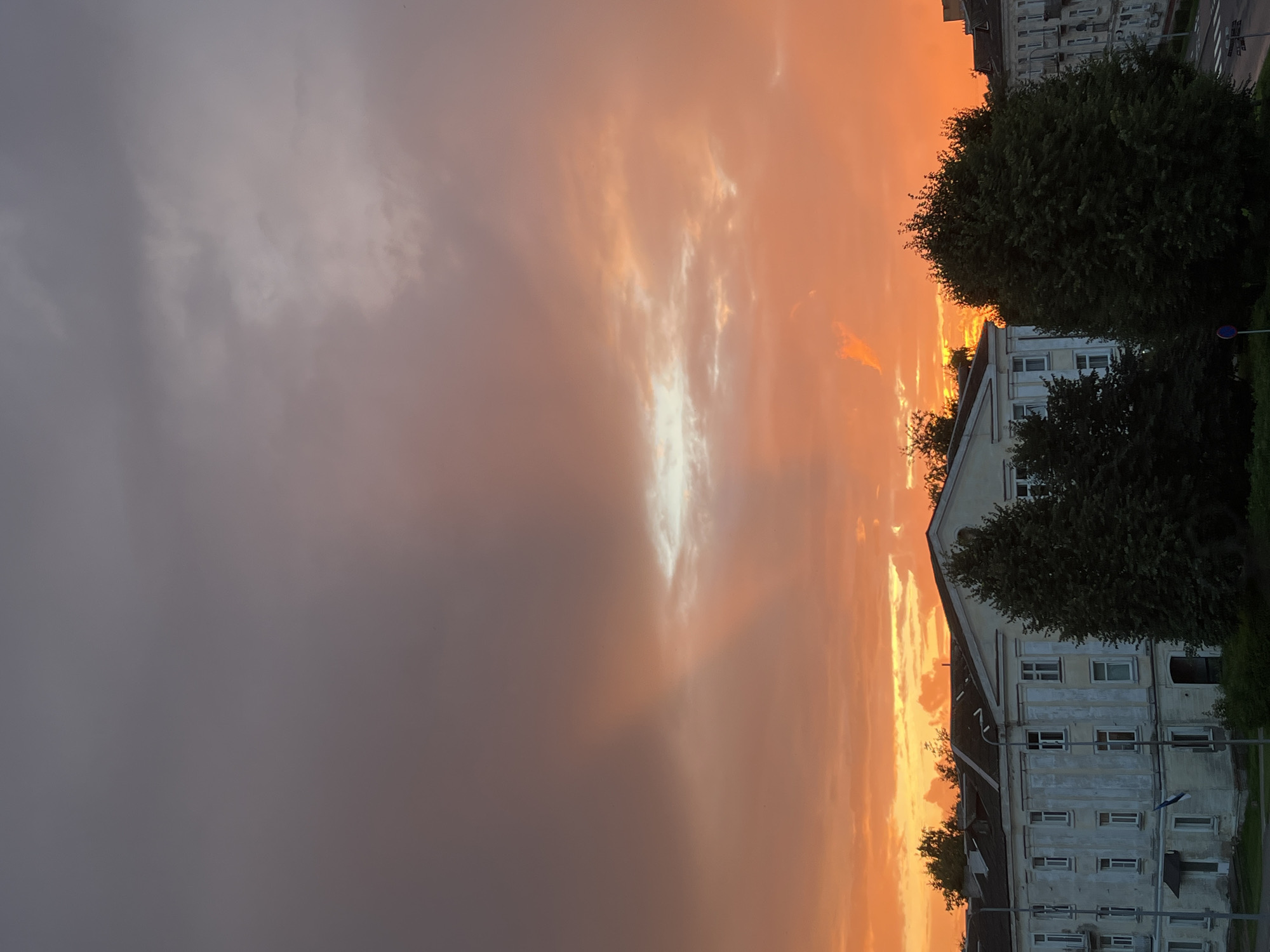 A panoramic, eye-level, outdoor, dusk photograph captures a dramatic sky over the city of Kohtla-Järve, Estonia. The sky is dominated by an intense orange and red sunset, with streaks of gray and purple clouds higher up. A bright, white patch of light breaks through the clouds in the center, creating a beam of light.
In the foreground, a long, white, multi-story building with numerous windows and a sloped, dark roof is visible. The facade of the building appears weathered. Several dark green, leafy trees are positioned in front of and to the right of the building, partially obscuring it. A small Estonian flag, blue and black, is visible on a flagpole attached to the building. Streetlights and a street sign are also present. To the right, another building, partly obscured by trees, is visible. The overall scene depicts an urban environment at sunset.