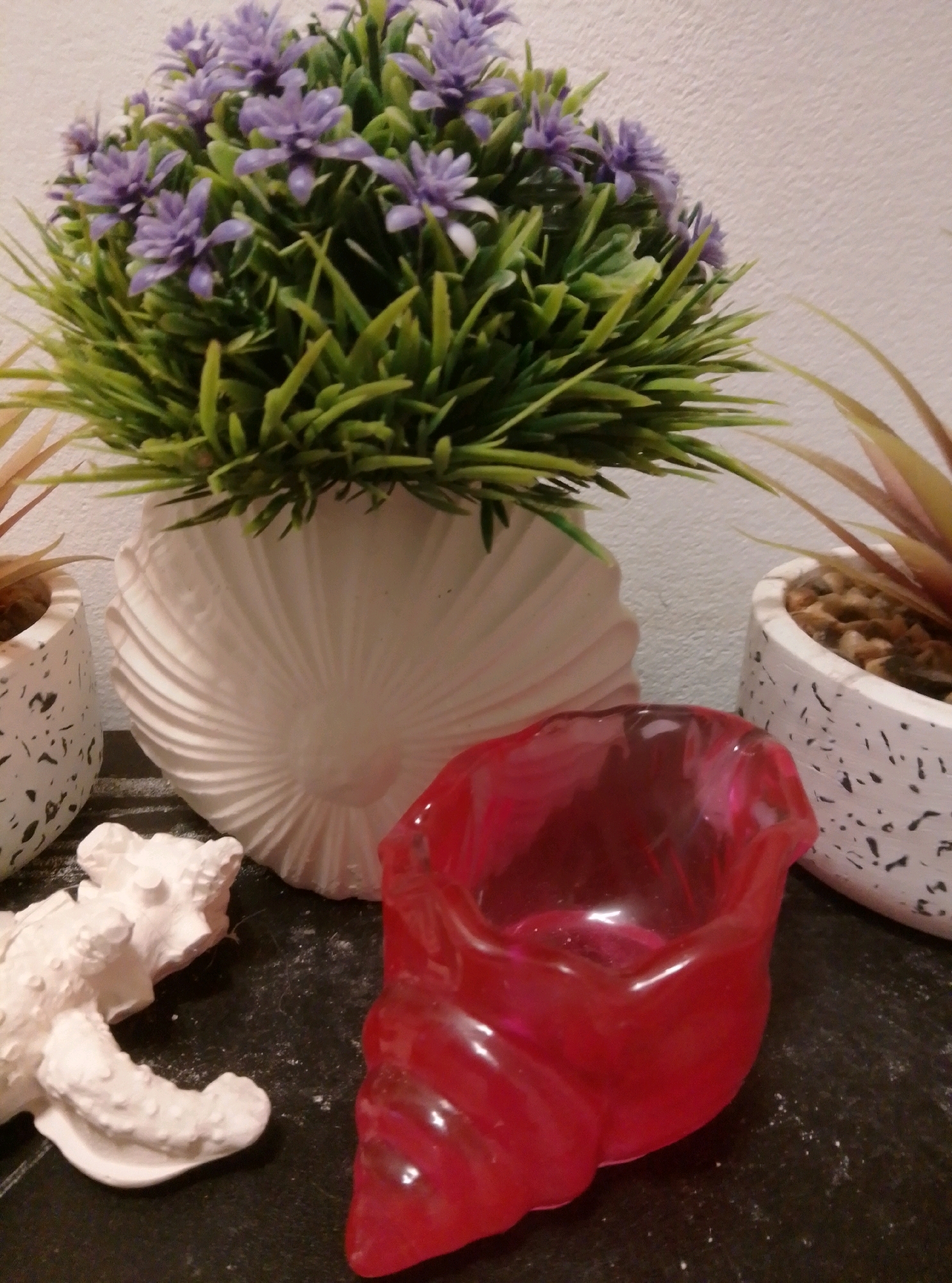 The image shows a still life arrangement on a dark surface. In the center is a white vase shaped like a scallop shell, holding a bouquet of artificial green foliage with small purple flowers. To the left of the vase, partially visible, is a small white, textured object resembling a piece of coral or a decorative figurine. To the right of the vase, and closer to the foreground, is a translucent red object with a glossy finish, also shaped like a shell or a swirl. In the background, to the left, is a white speckled planter with a plant featuring thin, reddish-green leaves. To the right, a similar white speckled planter holds a plant with spiky, light green and tan leaves. The background wall is textured and light-colored. No people or actions are depicted. The scene does not provide any information about the city of Wedel, Germany.