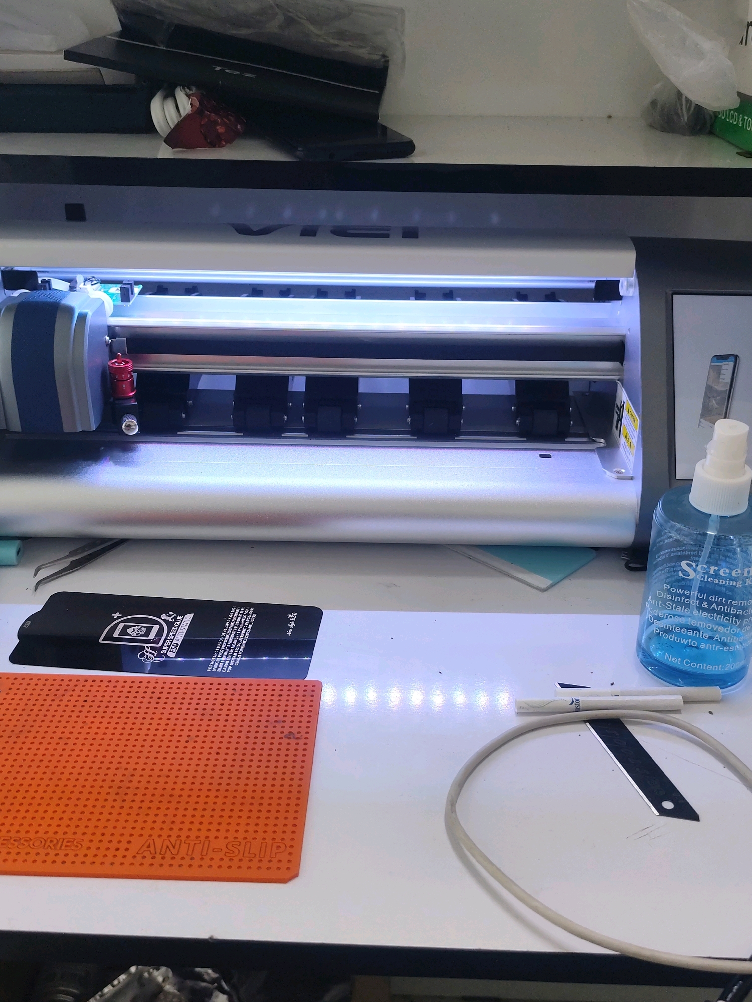 The image depicts a white cutting machine in what appears to be a workshop or electronics repair setting. On the machine's surface, various tools and supplies are laid out, including a bottle of "Screen Cleaning Kit" spray, tweezers, a black rectangular object with text and graphics that reads "SUPER SPEED GLUE", and an orange anti-slip mat. A thin cable and a razor blade are also visible on the table. The background shows shelves with some items, including what looks like electronic devices and packaging. The overall environment is lit by artificial lights, with a blueish glow emanating from the cutting machine. No people are visible in the image. The location of Kyzyltepa, Uzbekistan, is not discernible from the visual information provided in the image.