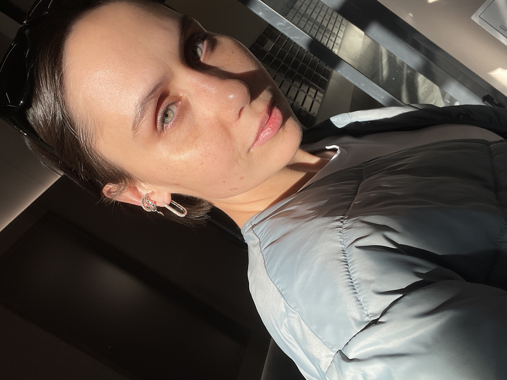 The image is a selfie of a young woman with fair skin, light-green eyes, and dark hair pulled back. She is wearing silver earrings and a light-blue puffer jacket over a white t-shirt. Sunglasses are perched on top of her head. The lighting is bright and sunny, casting shadows across her face and the surrounding environment.
The background is partially obscured by shadows, but it appears to be an urban environment with a metal grate-like structure in the upper portion of the frame. No details suggest the location is specifically Pervomaysky, Russia.