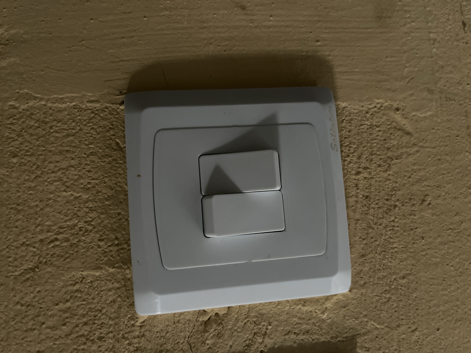 A white double-gang light switch is mounted on a textured, yellowish-brown wall. The switch plate is rectangular with rounded corners, and the two rocker switches are positioned side-by-side in the center. The left switch appears to be in the down position, while the right switch is in the up position. A small, faint logo, possibly a brand name, is visible on the lower right corner of the switch plate. The wall surface is rough and uneven, with visible cracks and irregularities. The lighting suggests an indoor environment.