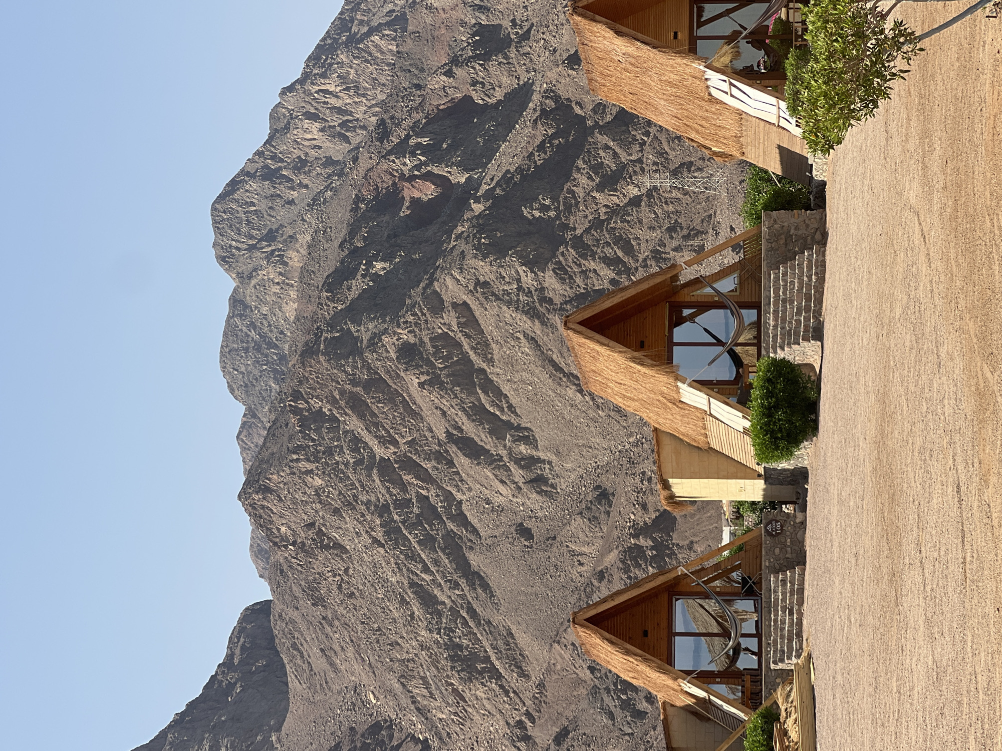 Three A-frame cabins with thatched roofs are arranged in a row on a sandy surface in front of a rocky mountain range under a clear blue sky. The cabins are made of wood and feature large glass windows and doors, with hammocks visible inside the first and third cabins. Stone steps lead up to the entrance of the first cabin, which also has a small stone wall and a sign with the number "105". Green bushes are planted in front of the cabins. The mountains in the background are rugged and dry, with visible rock formations and a power pylon in the distance.