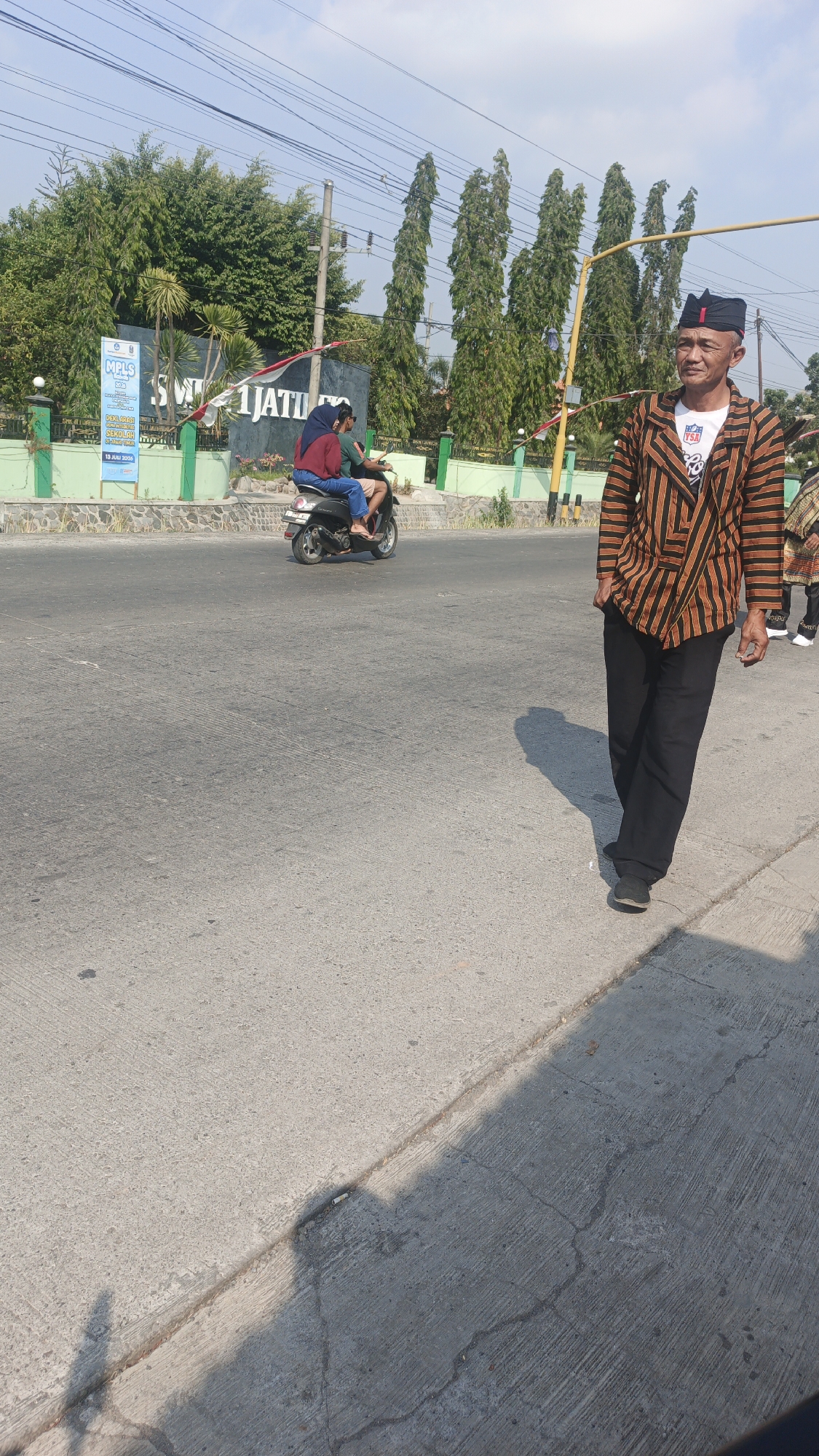 In Sumengko, Indonesia, a man is walking on the right side of the image. He is wearing a black and orange striped jacket, black pants, and a black cap with red trim. He is looking towards the left side of the image. Behind him, a scooter is driving away from the camera, carrying two passengers. In the background, there is a building with the sign "SMK 1 JATIM" and several tall green trees. The sky is blue with some clouds. The scene appears to be on a sunny day.