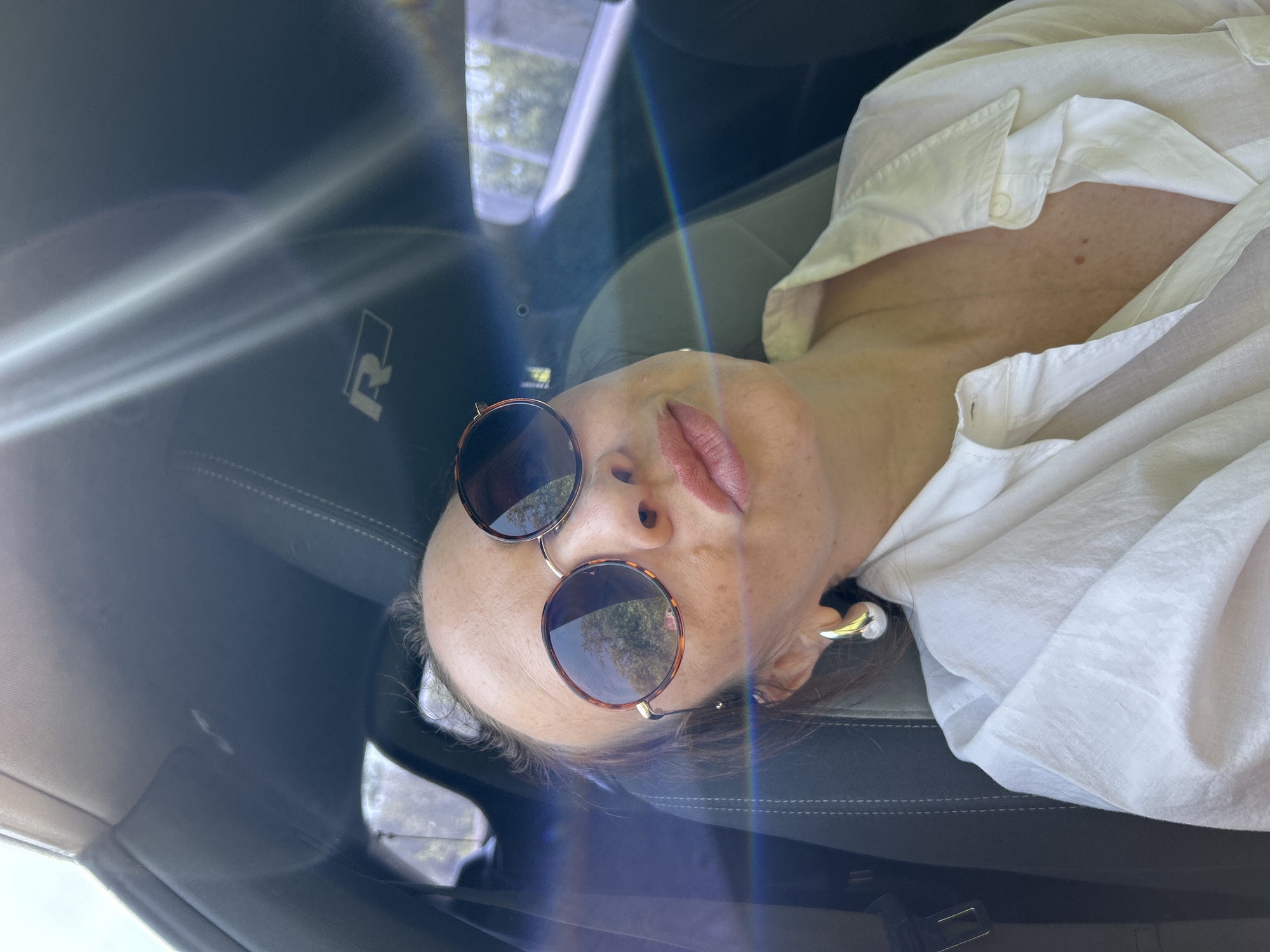 The image is an interior view of a car, likely taken from the passenger seat. A woman wearing round sunglasses and a white button-down shirt is reclining against the seat. The car appears to be parked, and some foliage can be seen through the car windows. The seat has a logo of "R". The exact location of the city Votkinsk, Russia, cannot be determined from the image.